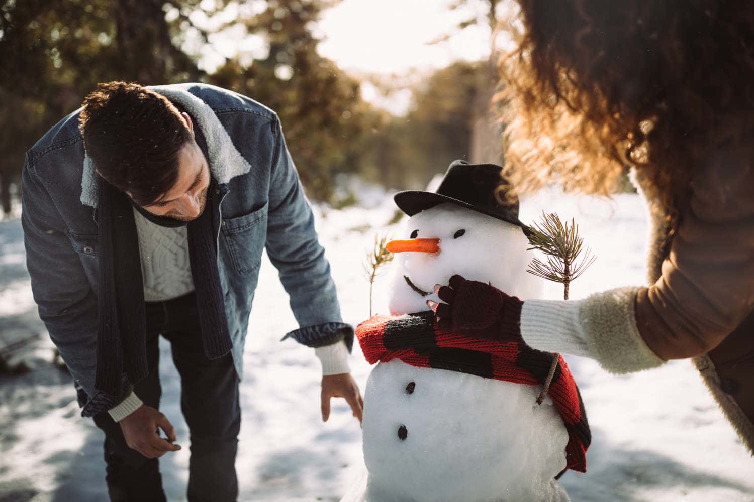 雪人 桌布 壁紙 — 一對情侶穿著保暖的冬季服裝，在陽光明媚的雪地森林中，輕輕地用圍巾和帽子裝飾雪人。