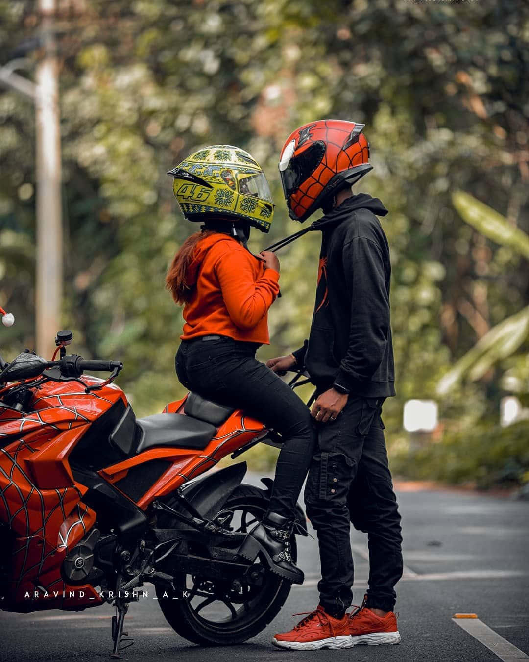 biker 桌布 壁紙 — 一對情侶穿著相配的紅黑蜘蛛主題頭盔和裝備，在綠樹成蔭的道路旁，於一輛充滿活力的橙色機車旁邊分享親密時刻。