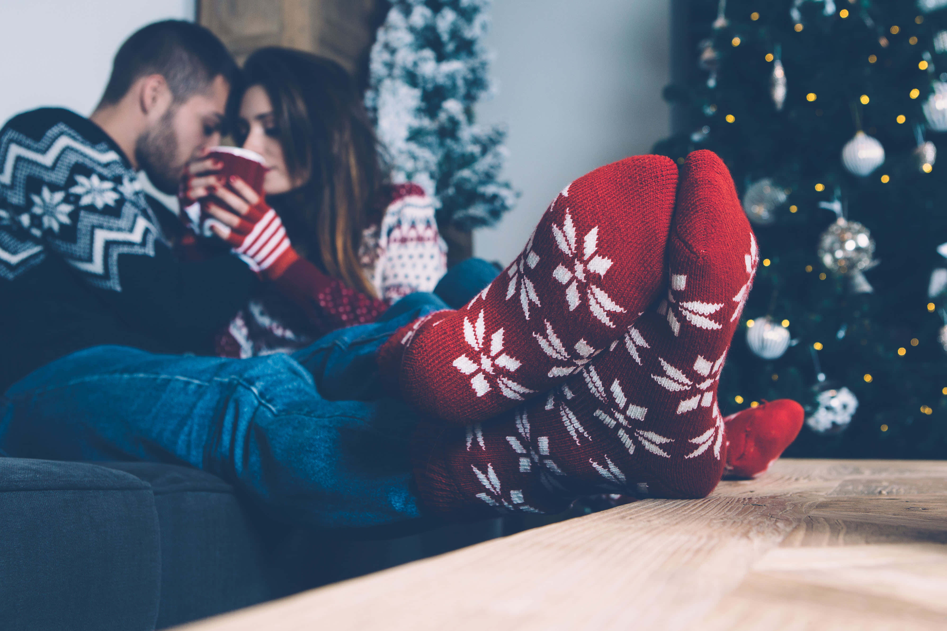 Couples Christmas 桌布 壁紙 — 一對情侶穿著節日毛衣和紅色雪花襪，在閃爍的聖誕樹旁的沙發上依偎。