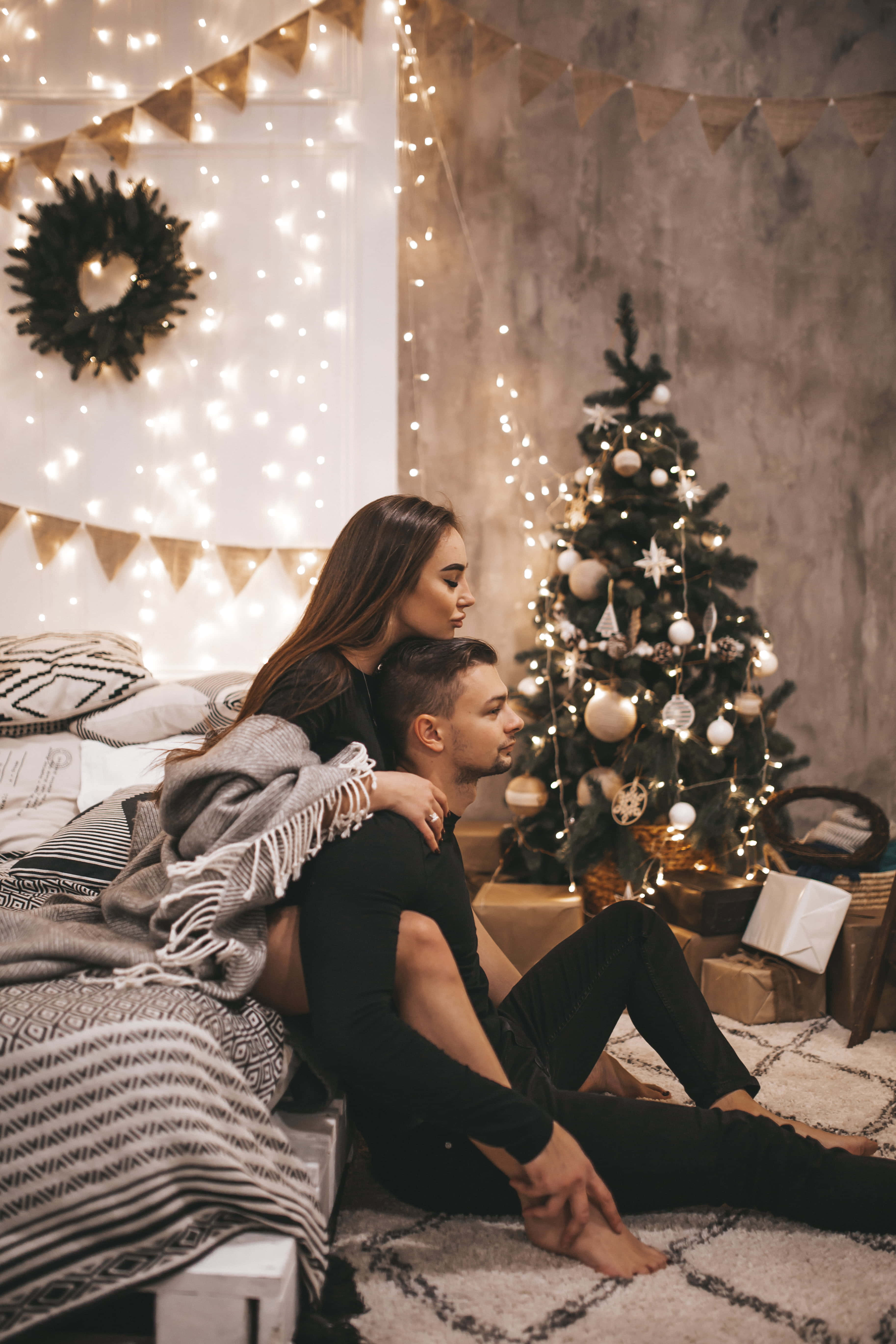 Couples Christmas 桌布 壁紙 — 一對情侶舒適地坐在圖案地毯上，旁邊是一棵裝飾著仙女燈和粗麻布橫幅的聖誕樹。