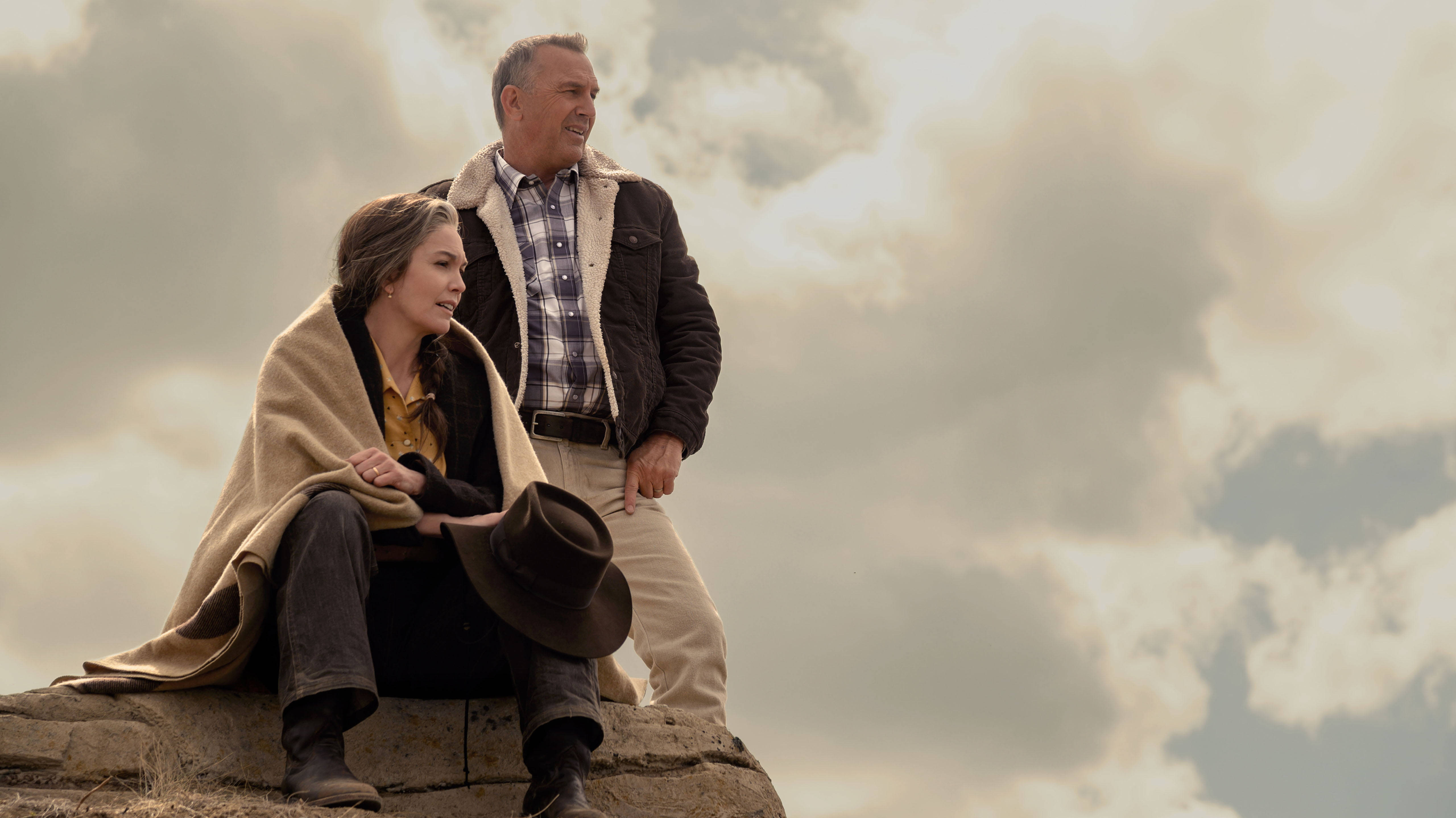 Kevin Costner 桌布 壁紙 — 一對情侶裹著毯子坐在岩石露頭上，在廣闊多雲的天空下分享寧靜的時刻。