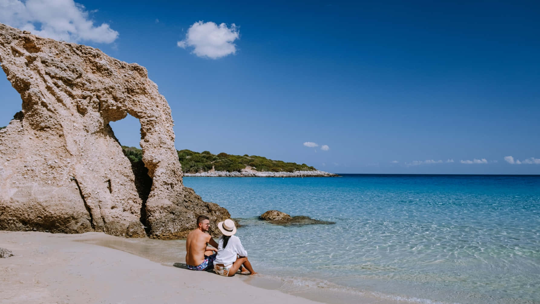 Greek Beach 桌布 壁紙 — 一對情侶親密地坐在陽光明媚的海灘上，旁邊是一個巨大的天然岩石拱門，俯瞰著清澈藍天下的碧綠海水。
