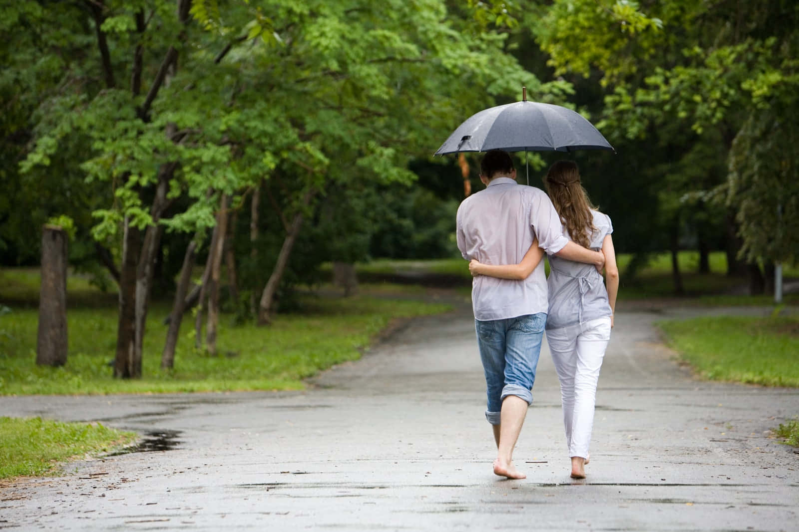 情侶 桌布 壁紙 — 一對情侶赤腳撐傘走在潮濕的公園小徑上，周圍是茂密的綠樹。