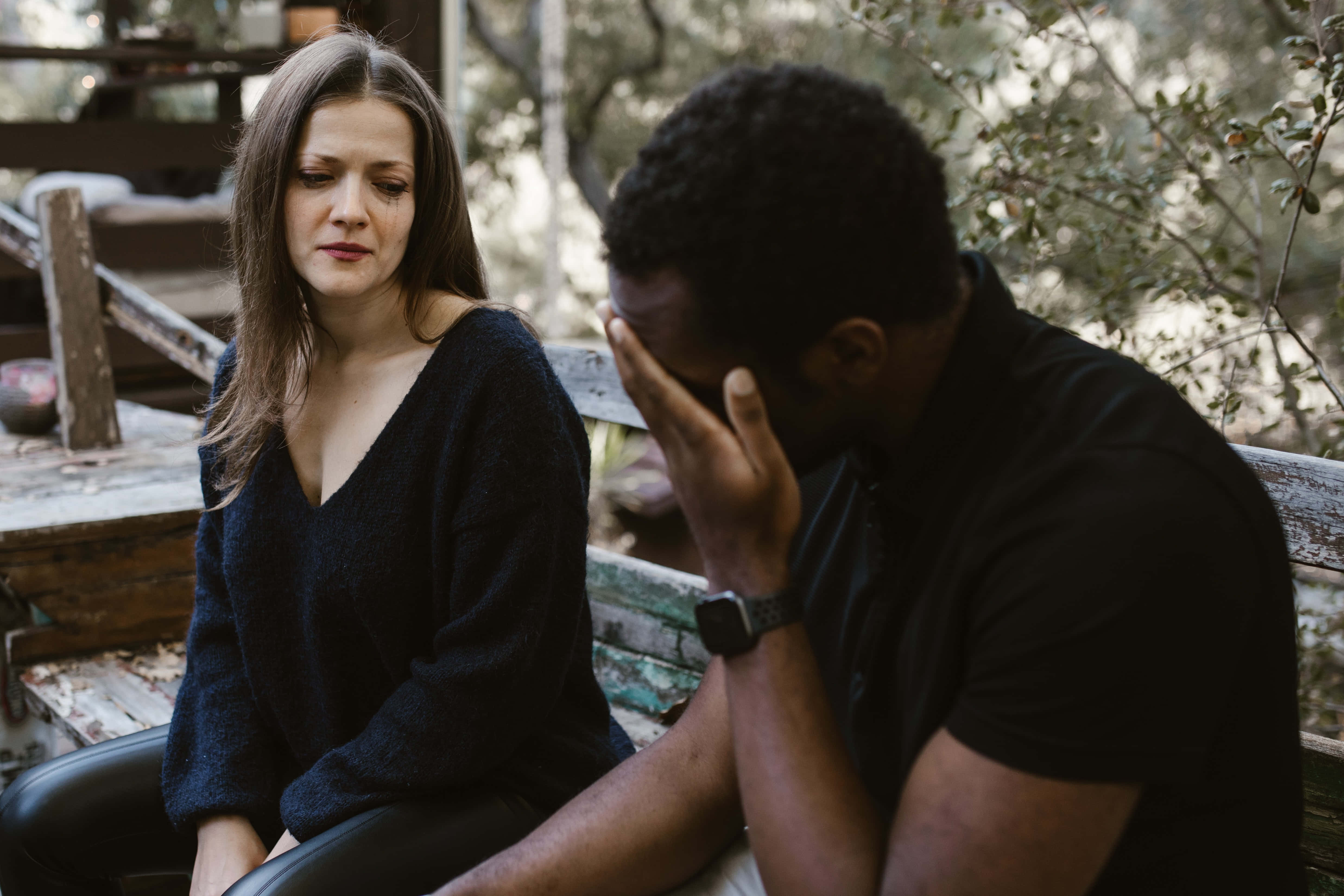 Sad Couple 桌布 壁紙 — 一對憂鬱的情侶坐在森林中一張風化的木長椅上，男人痛苦地掩面，而女人則帶著沉思的表情看著他。