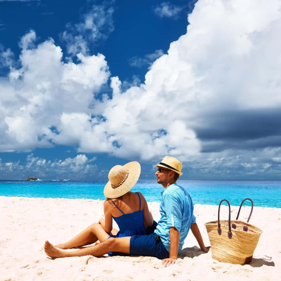 sand 桌布 壁紙 — 一對戴草帽的情侶坐在白色沙灘上，面向碧綠海洋，天空蔚藍，白雲朵朵。