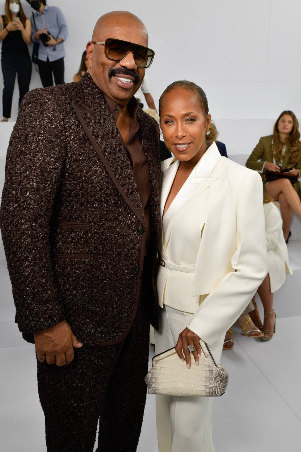 steve harvey 桌布 壁紙 — 一對時尚情侶在時裝秀上自信地擺姿勢，男士身穿質感棕色西裝和太陽眼鏡，女士身穿純白西裝，手持鱷魚紋手提包。