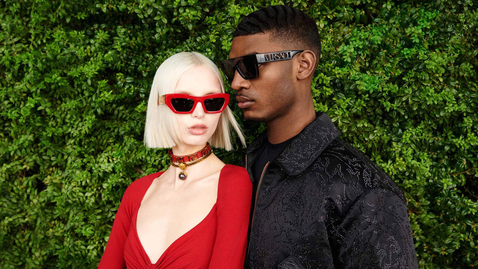 Sunglasses 桌布 壁紙 — 一對時尚情侶在茂密的綠色籬笆前擺姿勢，戴著大膽的紅色和黑色太陽眼鏡，穿著時尚服裝。