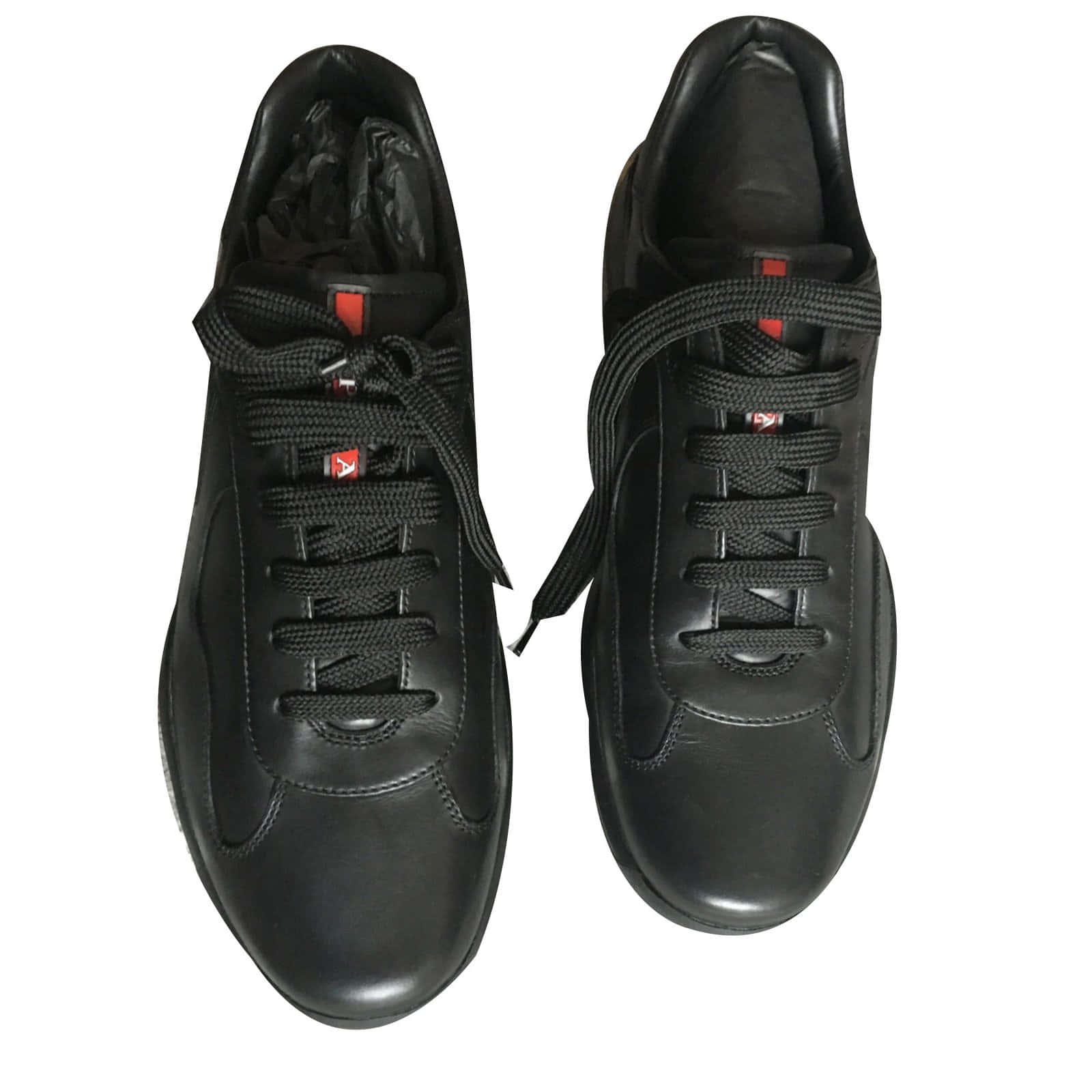 Prada 桌布 壁紙 — 一對時尚黑色皮革運動鞋，帶有紅色標誌點綴和黑色鞋帶，從上方看白色背景。