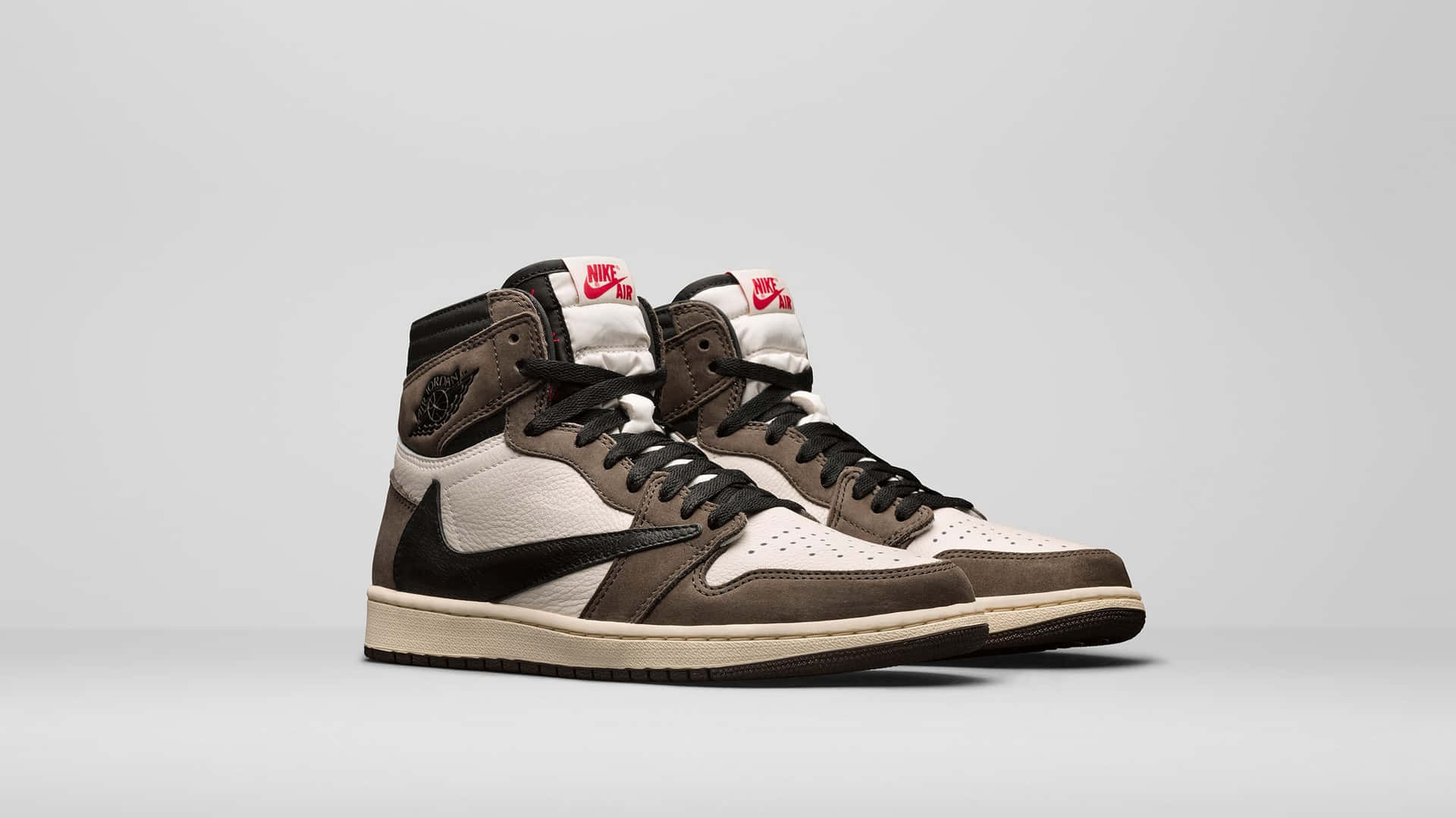 Travis Scott Jordan 1 桌布 壁紙 — 一對棕色和白色的高筒運動鞋，配有黑色鞋帶和紅色Nike品牌標誌，展示在純灰色背景上。