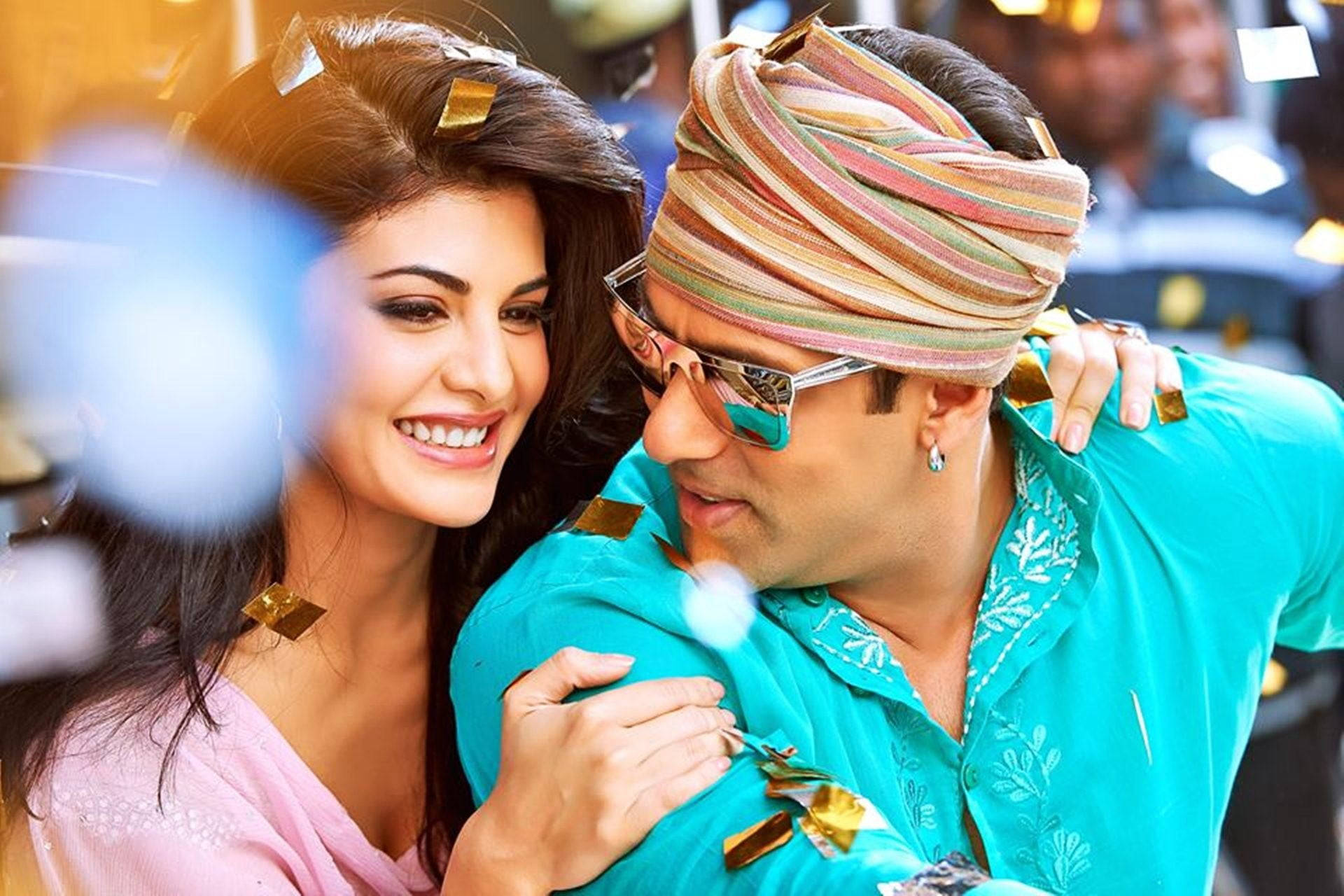 Bollywood HD 桌布 壁紙 — 一對歡樂的情侶穿著鮮豔的傳統服飾，在飄落的金色紙屑中分享嬉戲的時刻。