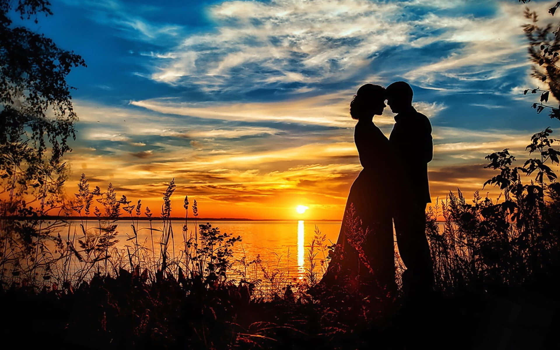 Romantic Love 桌布 壁紙 — 一對浪漫情侶在平靜湖面上方燦爛的日落映襯下的剪影，金色光芒在水面上反射，雲彩壯麗。