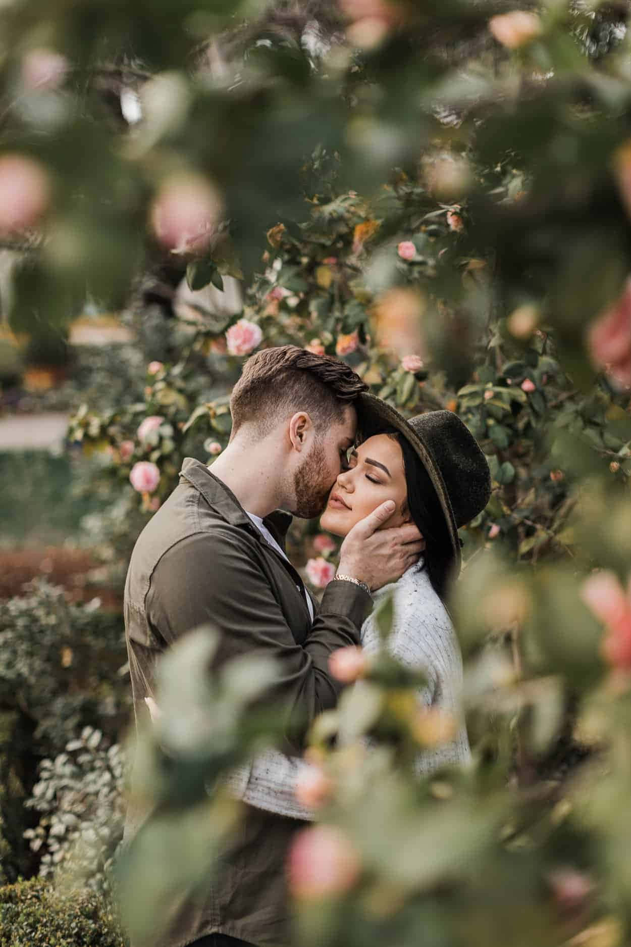 Beautiful Couple 桌布 壁紙 — 一對溫柔的情侶在茂密花園中，被柔焦的玫瑰叢框住，分享親密的親吻。