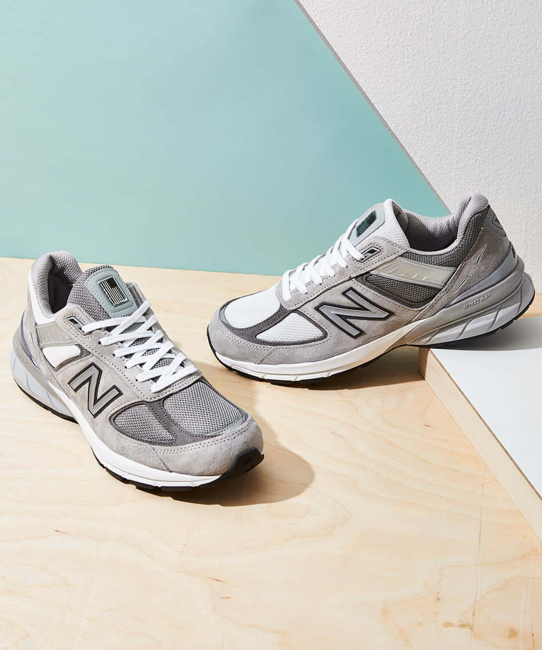 New Balance 桌布 壁紙 — 一對灰色New Balance運動鞋搭配白色鞋帶，放置在淺色木質表面上，背景是藍綠色和白色的幾何牆壁。