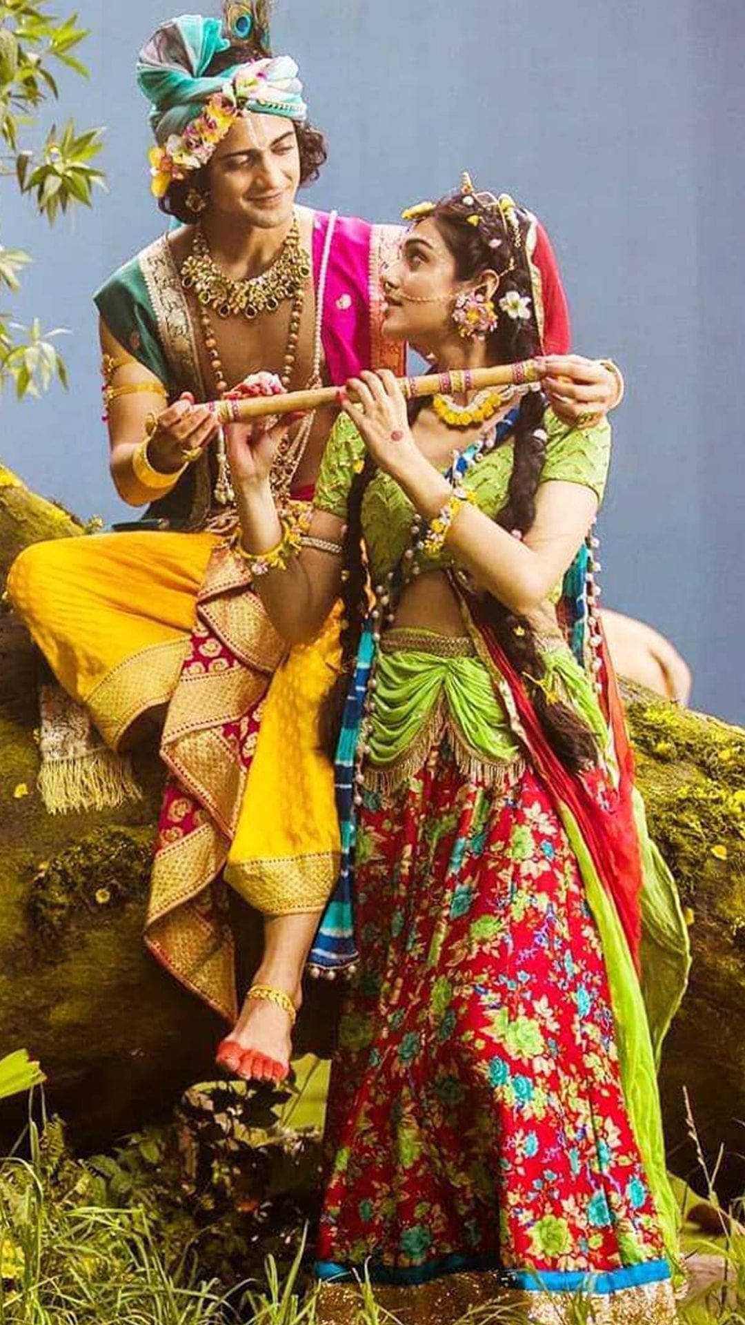 radha krishna serial 桌布 壁紙 — 一對男女穿著鮮豔的傳統印度服飾，在長滿苔蘚的木頭上分享歡樂時光，她吹著長笛，他微笑著在她身邊。