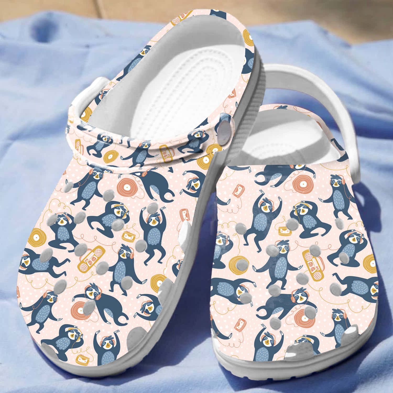Crocs 桌布 壁紙 — 一對白色木屐，帶有藍色和橙色的俏皮樹懶圖案，在明亮的陽光下放置在柔軟的藍色布料上。