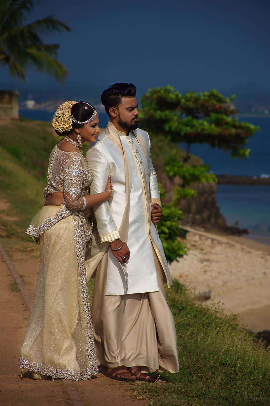 Wedding 桌布 壁紙 — 一對穿著傳統奶油色婚禮服飾的情侶站在海岸小徑上，俯瞰著清澈藍天下碧綠色的海洋。
