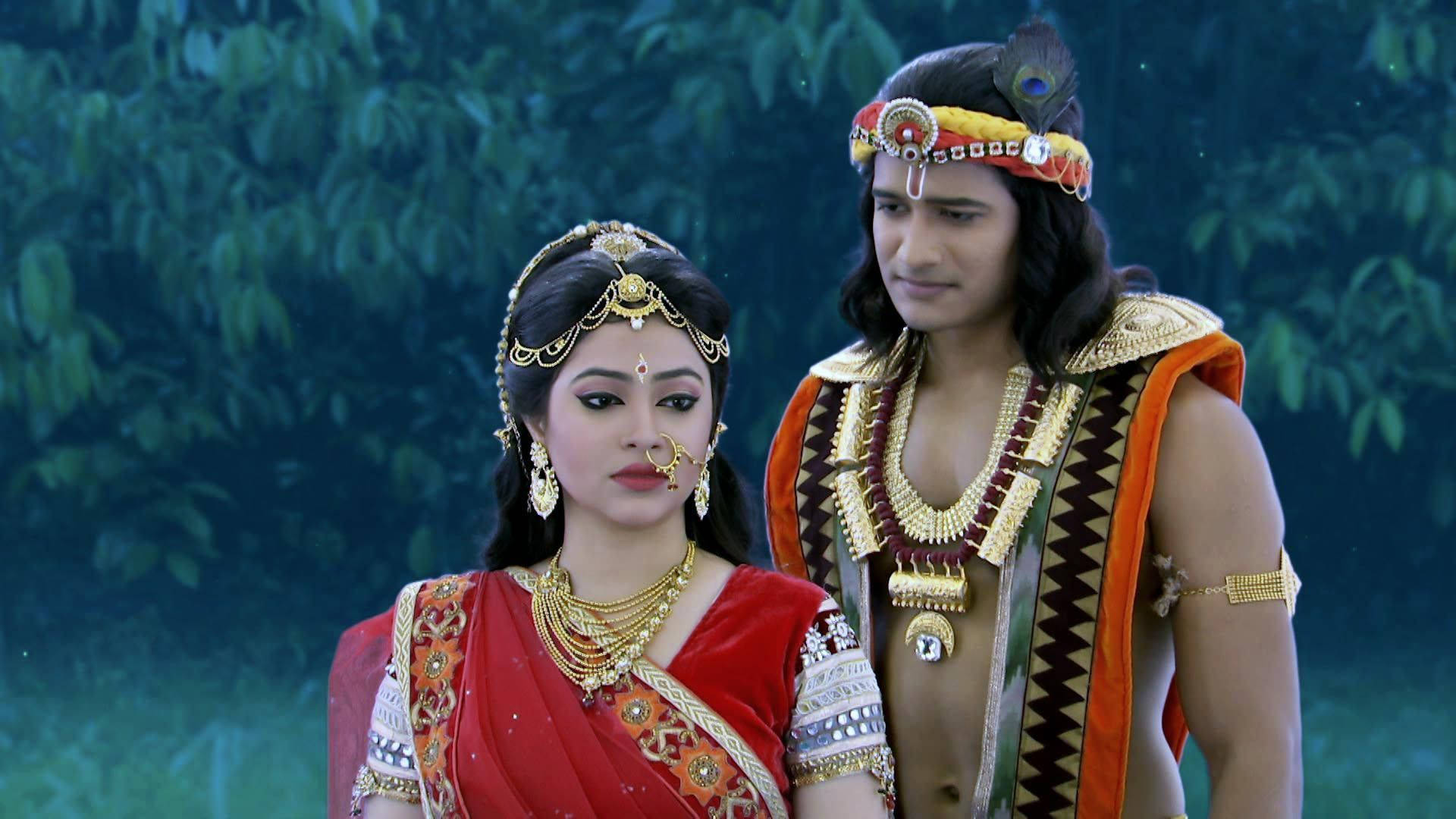 radha krishna serial 桌布 壁紙 — 一對穿著傳統服飾的印度夫婦，佩戴華麗的金飾，身穿鮮豔的紅色和橙色服裝，站在茂密的綠葉叢中。