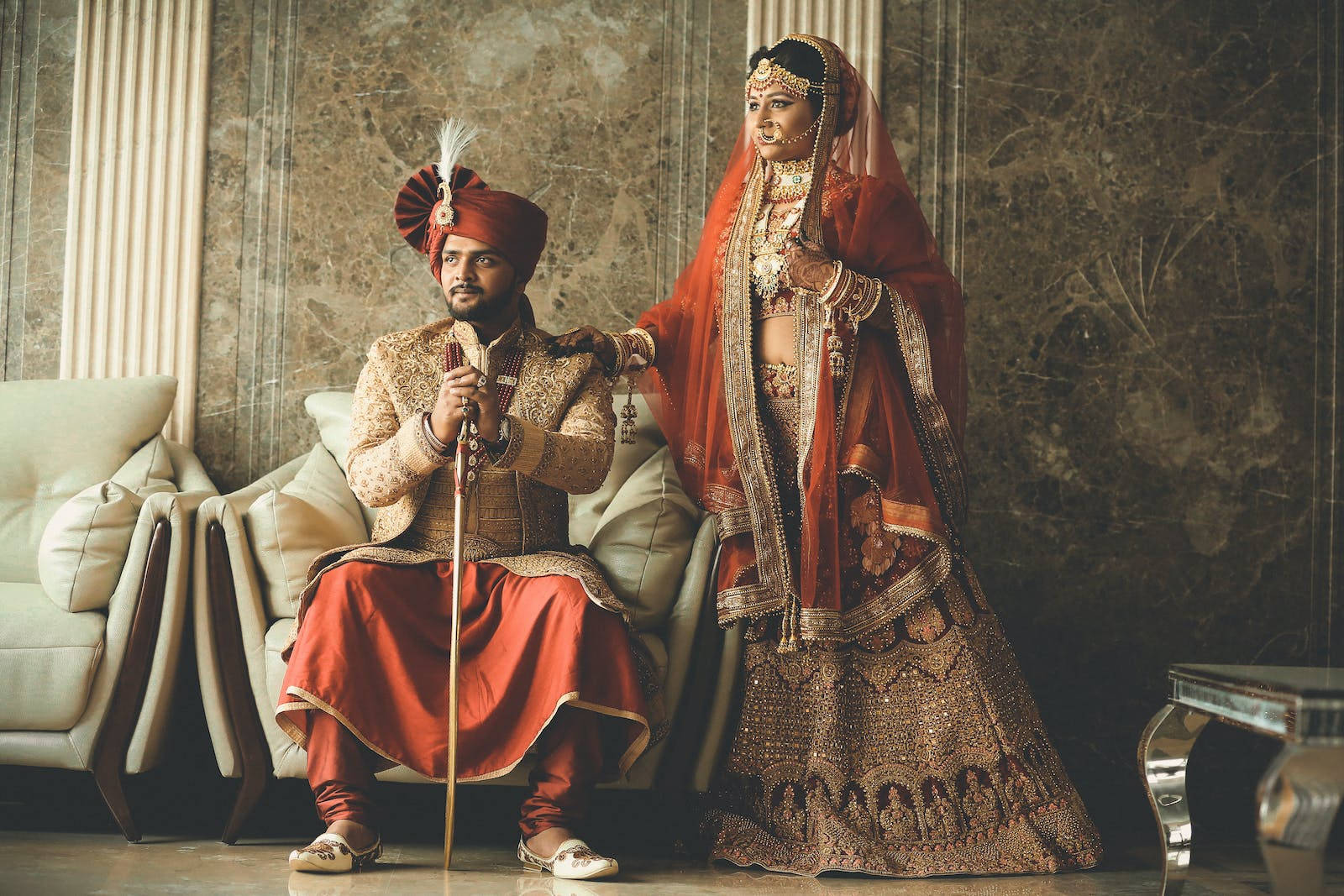 Indian 桌布 壁紙 — 一對穿著傳統服飾的印度夫婦，在雲石牆和米色沙發前，以華麗的婚禮服飾優雅地擺姿勢。