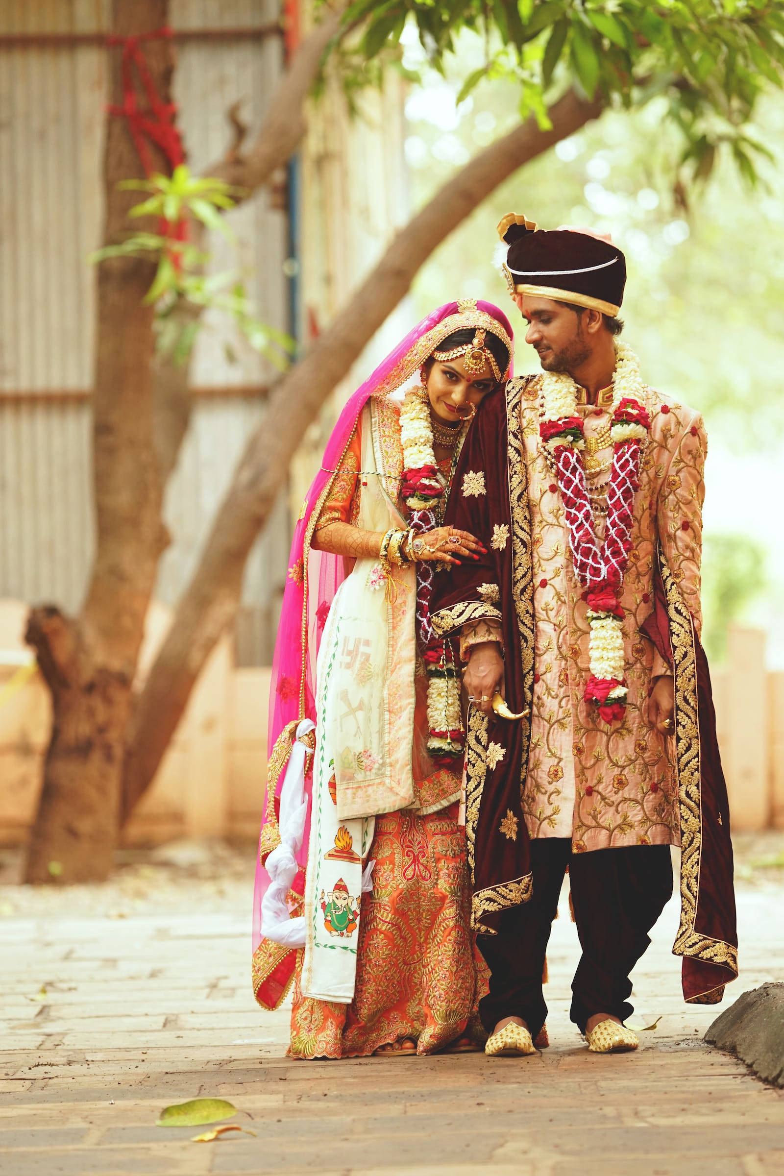 Indian 桌布 壁紙 — 一對穿著傳統服飾的印度新婚夫婦在陽光明媚的庭院中，手牽手走在枝葉茂密的樹下。