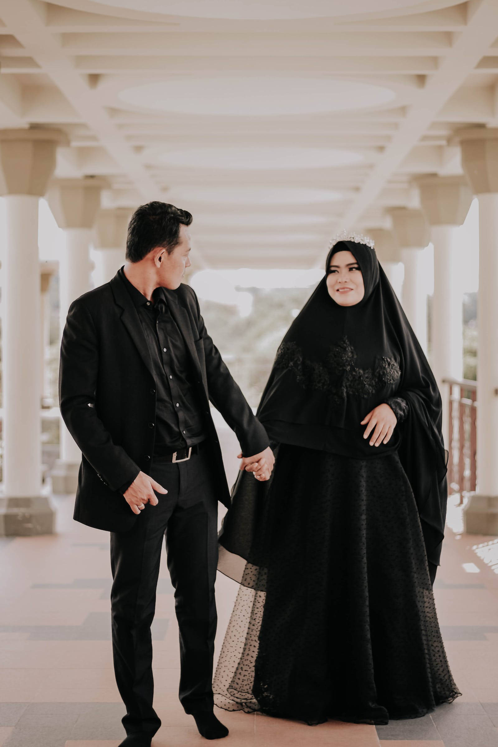 muslim 桌布 壁紙 — 一對穿著優雅黑色服裝的情侶在明亮的白色柱廊結構下牽手，柔和自然光線灑落。