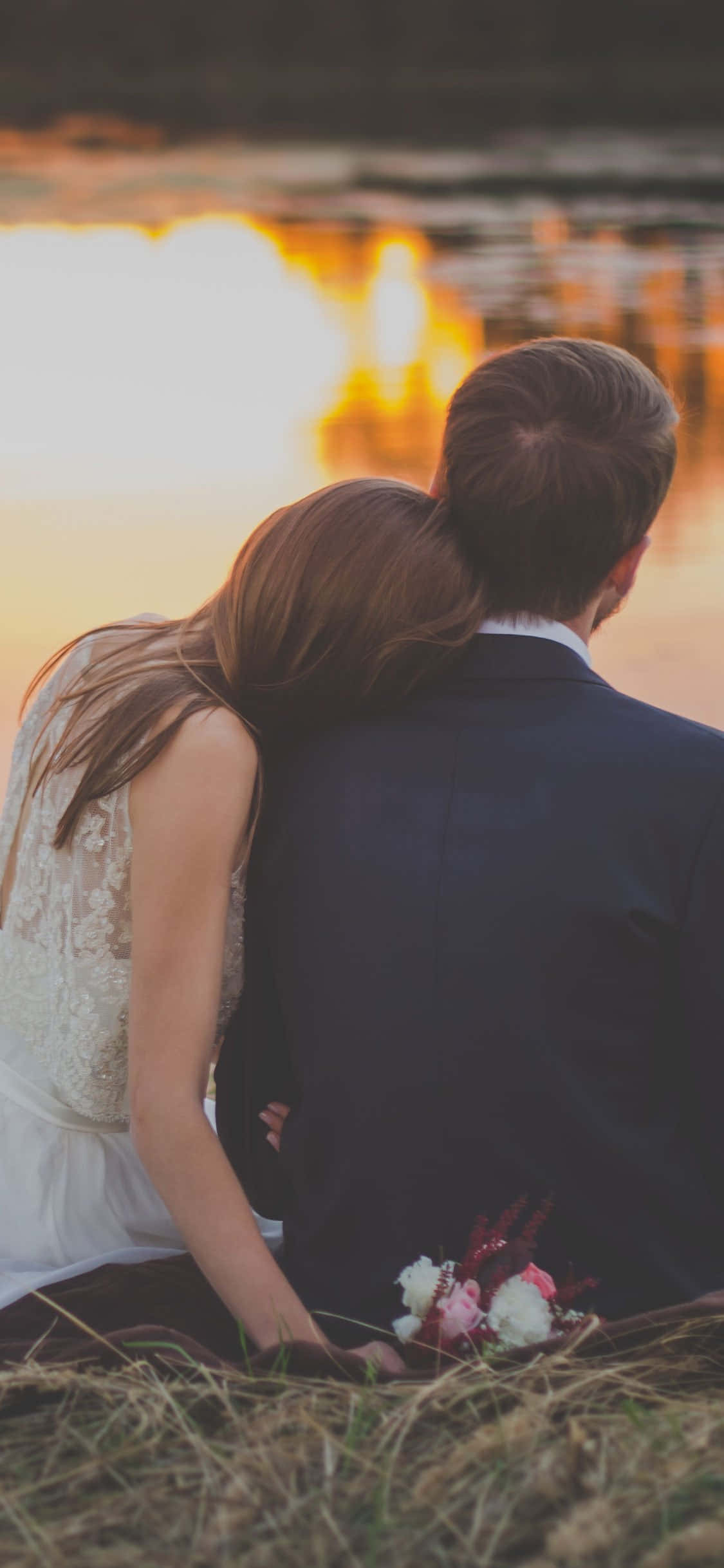 Wedding Couple 桌布 壁紙 — 一對穿著婚禮服飾的情侶背對背坐在草地上，觀賞水面上的金色日落。