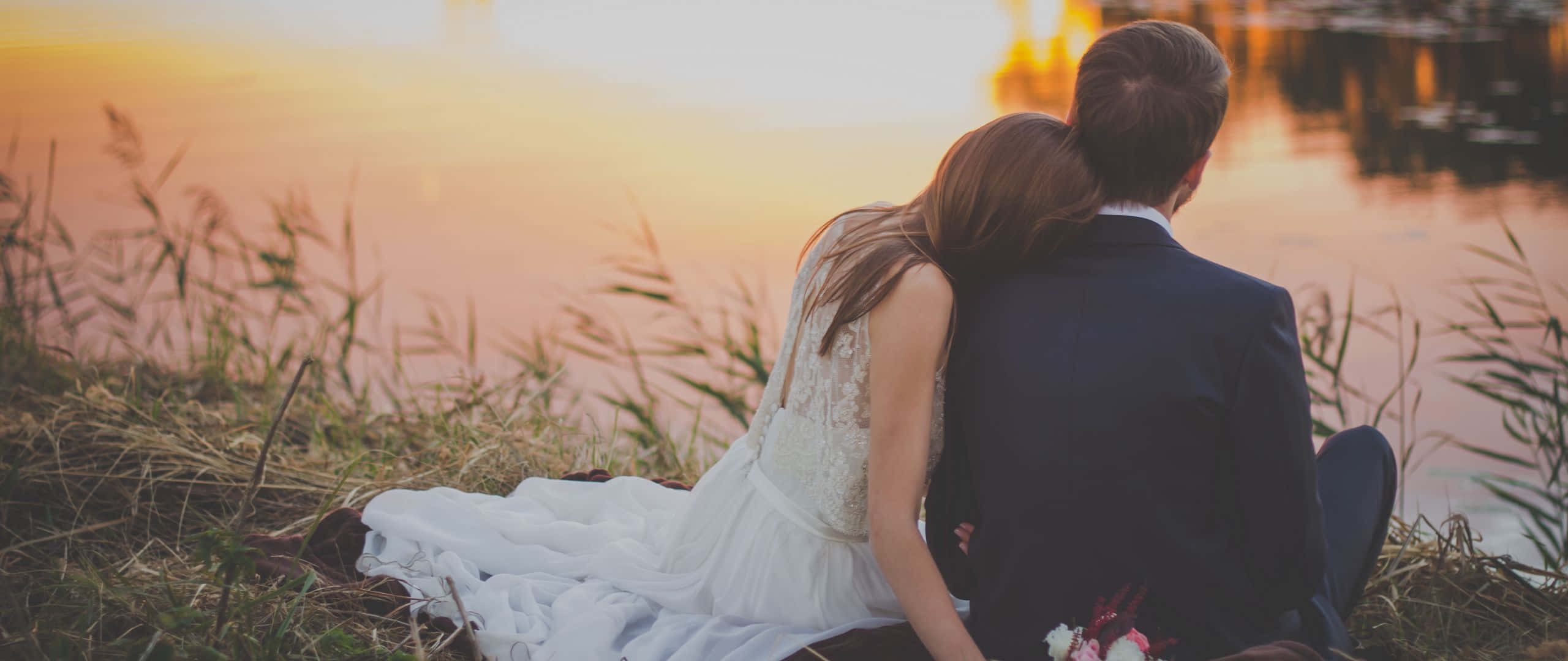 Wedding 桌布 壁紙 — 一對穿著婚禮服飾的情侶背對背坐在草地上，觀賞平靜水面上金色的日落。
