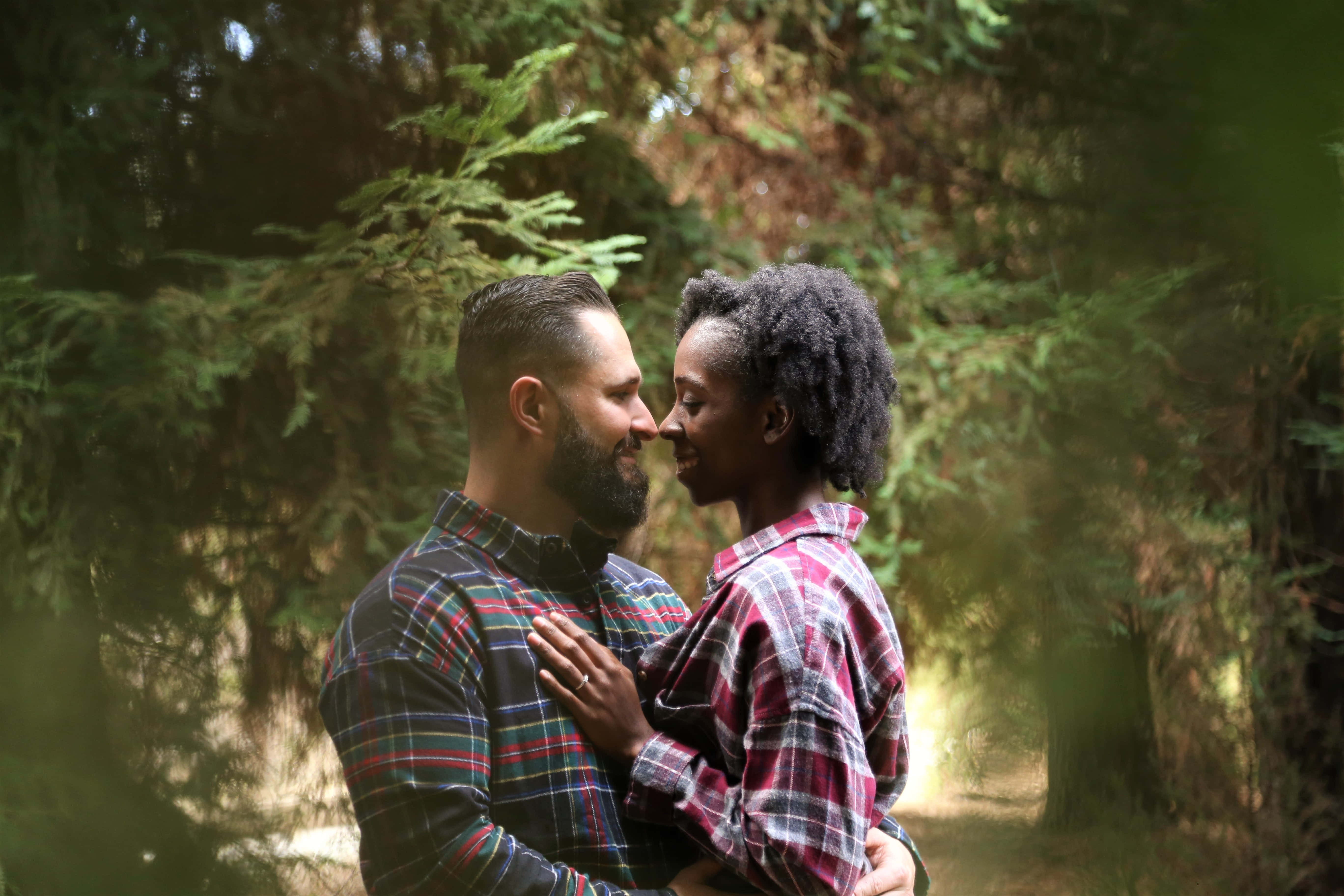Interracial 桌布 壁紙 — 一對穿著格紋襯衫的恩愛情侶，在陽光明媚的森林中，鼻尖對鼻尖，分享親密時刻，周圍是高聳的常綠樹。