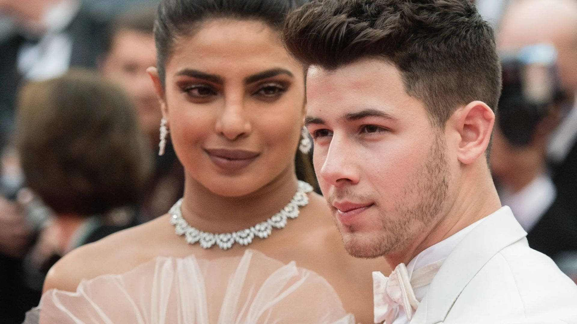 Nick Jonas 桌布 壁紙 — 一對穿著白色正裝的迷人夫婦在紅地毯活動上緊密擺姿勢，背景有閃爍的相機。