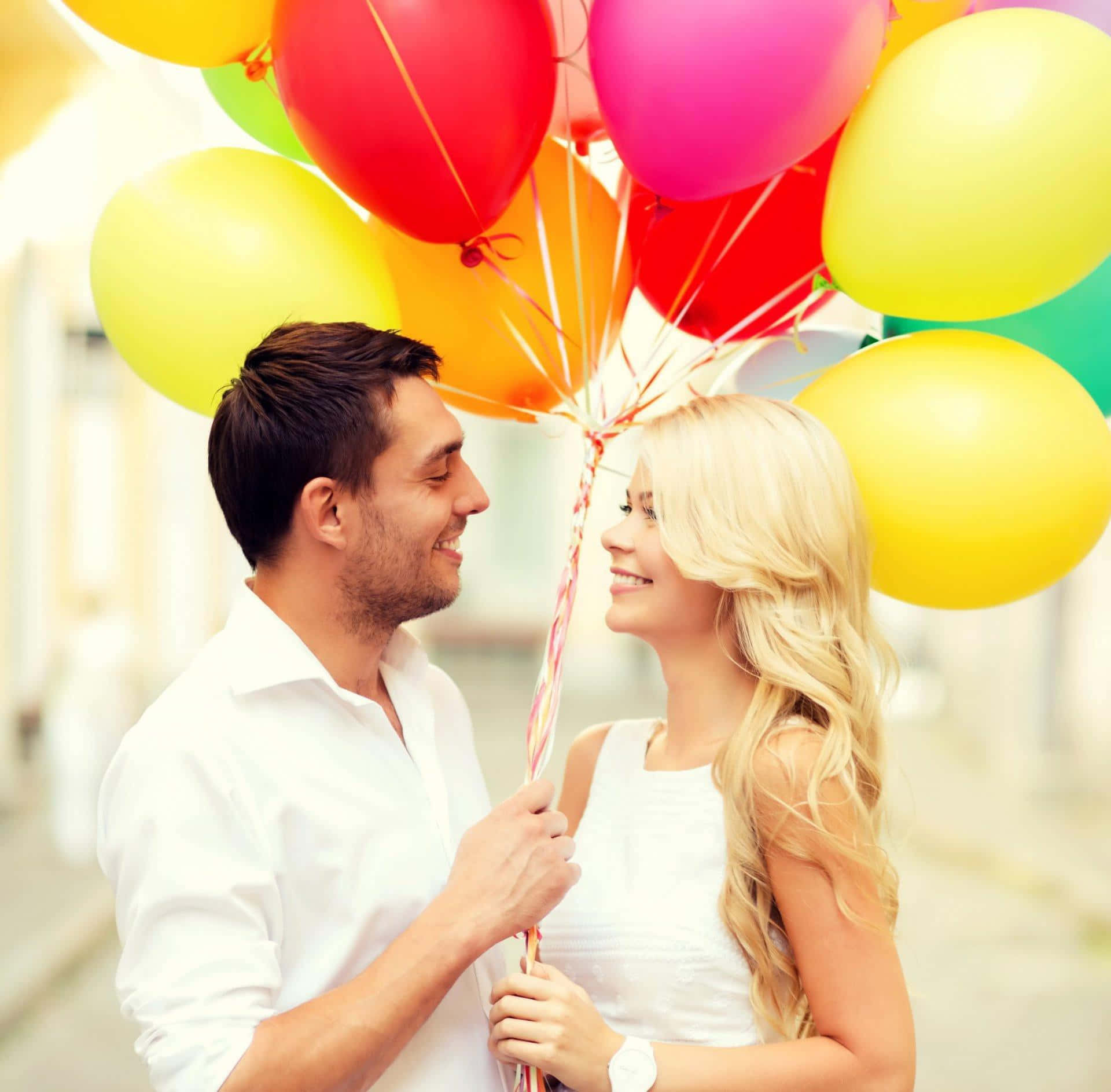 Balloons 桌布 壁紙 — 一對穿著白色衣服的微笑情侶在明亮的街道上分享歡樂時刻，手持一束彩色氣球。
