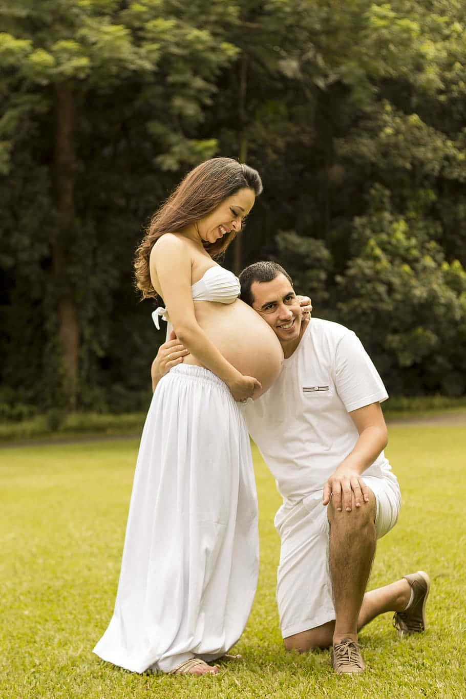Pregnant 桌布 壁紙 — 一對穿著白色衣服的快樂情侶在綠樹成蔭的草地上分享溫馨時刻。