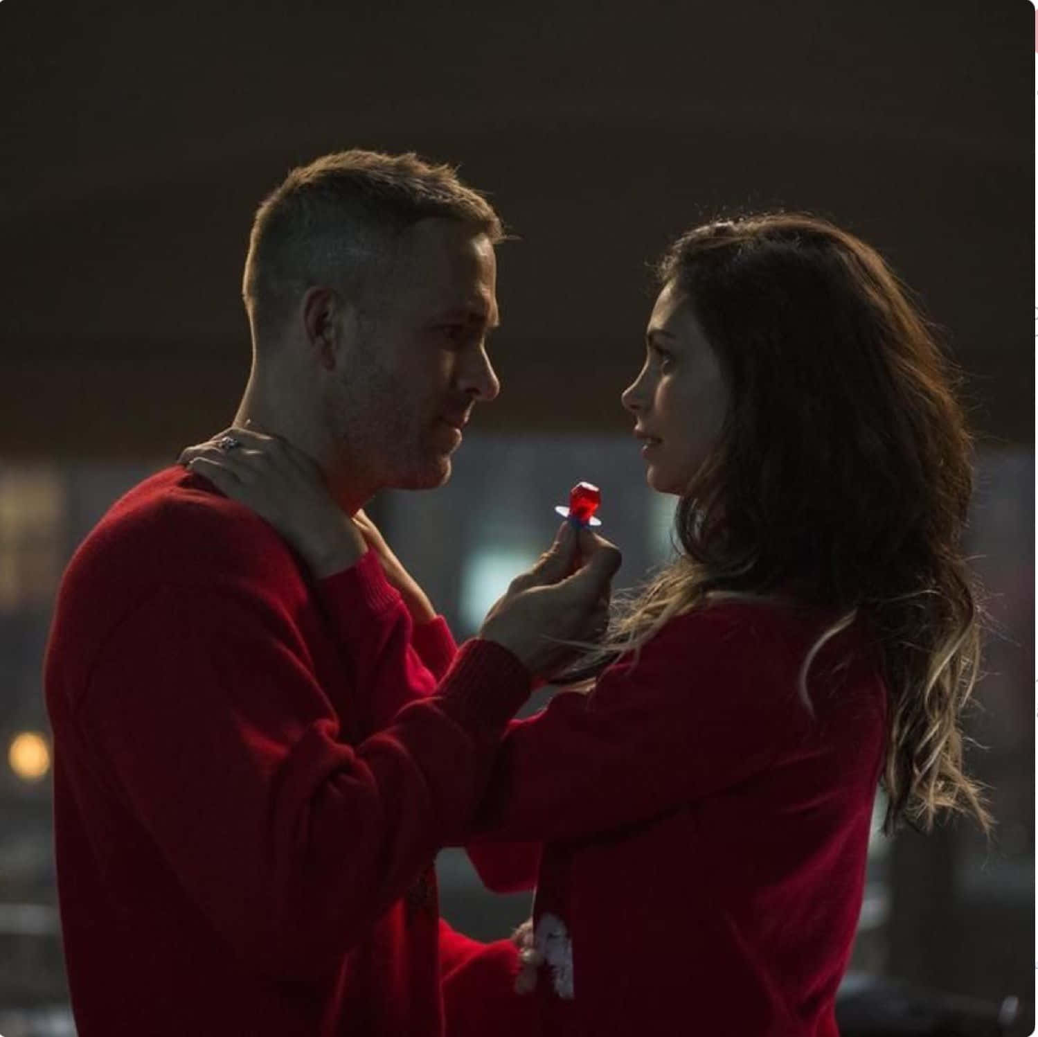 Deadpool And Vanessa 桌布 壁紙 — 一對穿著相配紅色毛衣的男女在昏暗燈光下分享親密時刻，她拿著一個紅色戒指盒面向他。