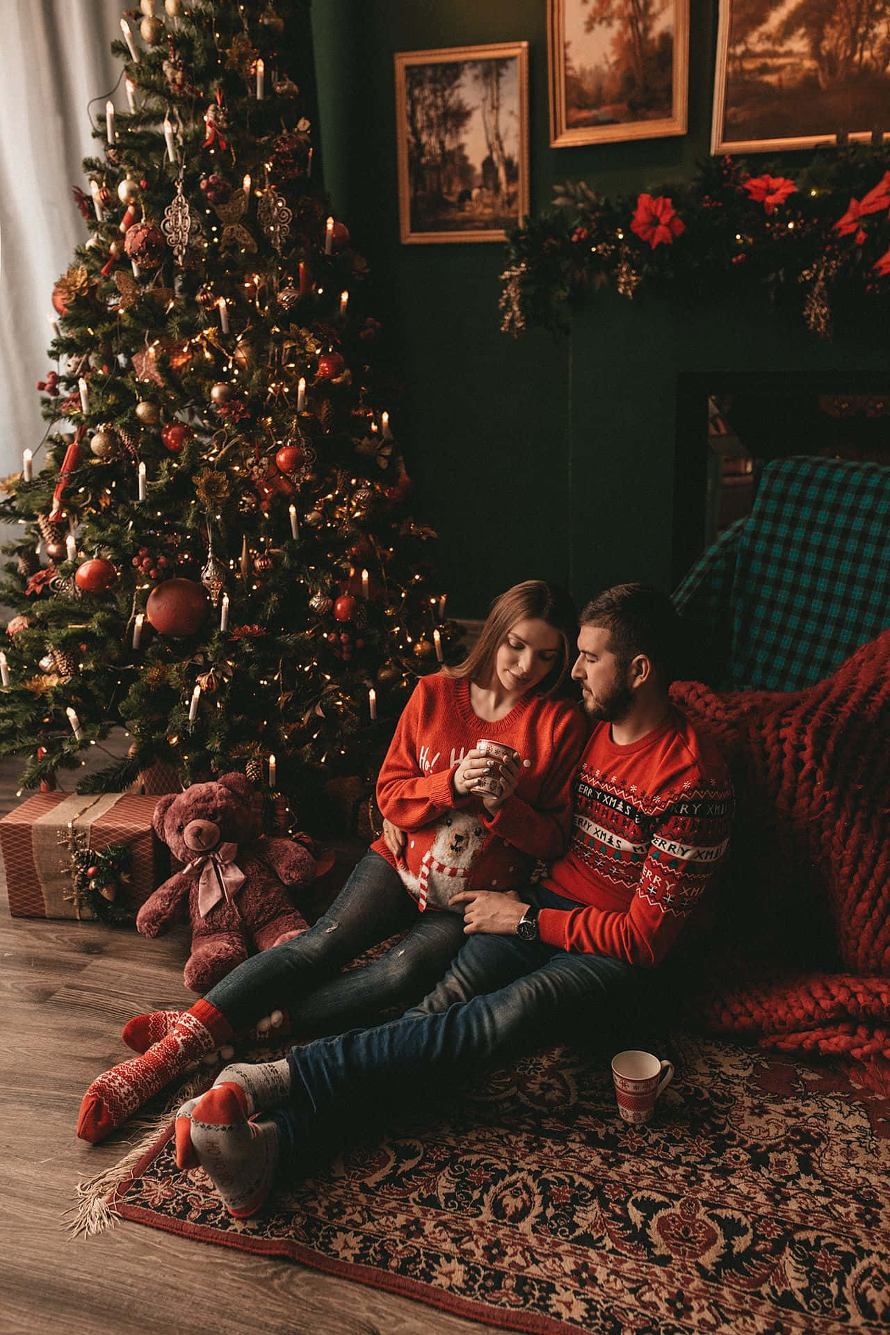 Couples Christmas 桌布 壁紙 — 一對穿著紅色毛衣的情侶在圖案地毯上擁抱一隻小狗，旁邊是裝飾著溫暖燈光的聖誕樹。