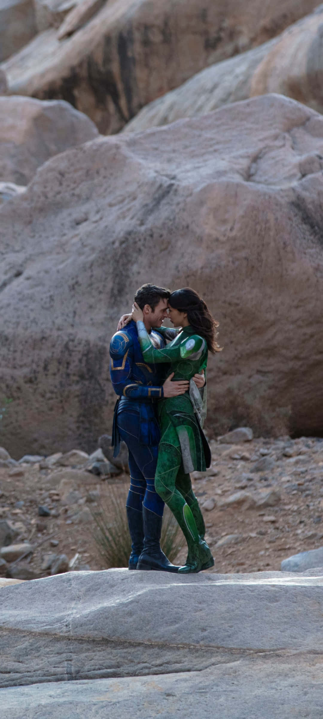 Eternals Movie 桌布 壁紙 — 一對穿著超級英雄服裝的情侶在溫暖陽光下的岩石沙漠景觀中溫柔擁抱。