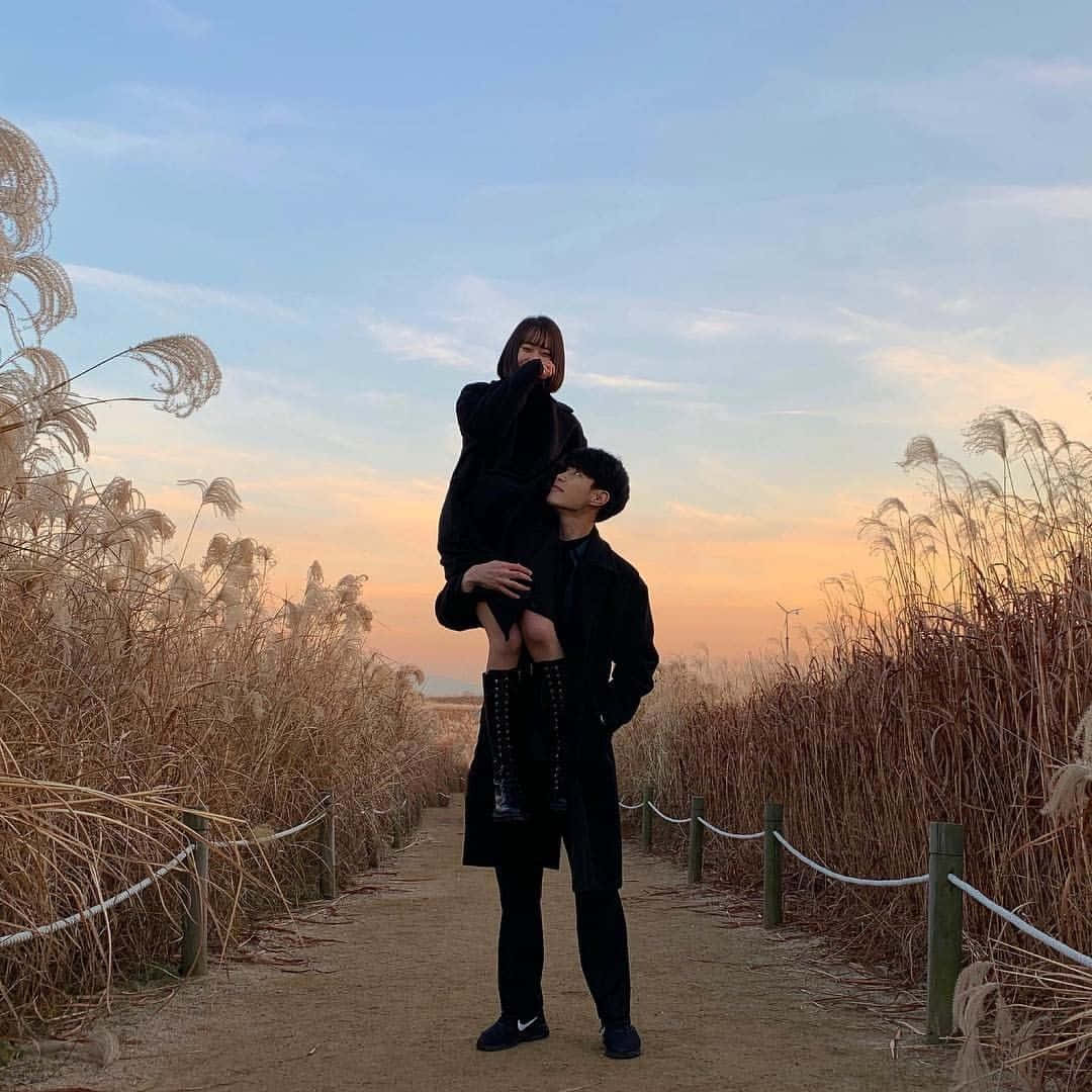 lover 桌布 壁紙 — 一對穿著黑色外套的情侶分享著溫柔的時刻，男人在夕陽天空下舉起女人，四周是高高的蘆葦。