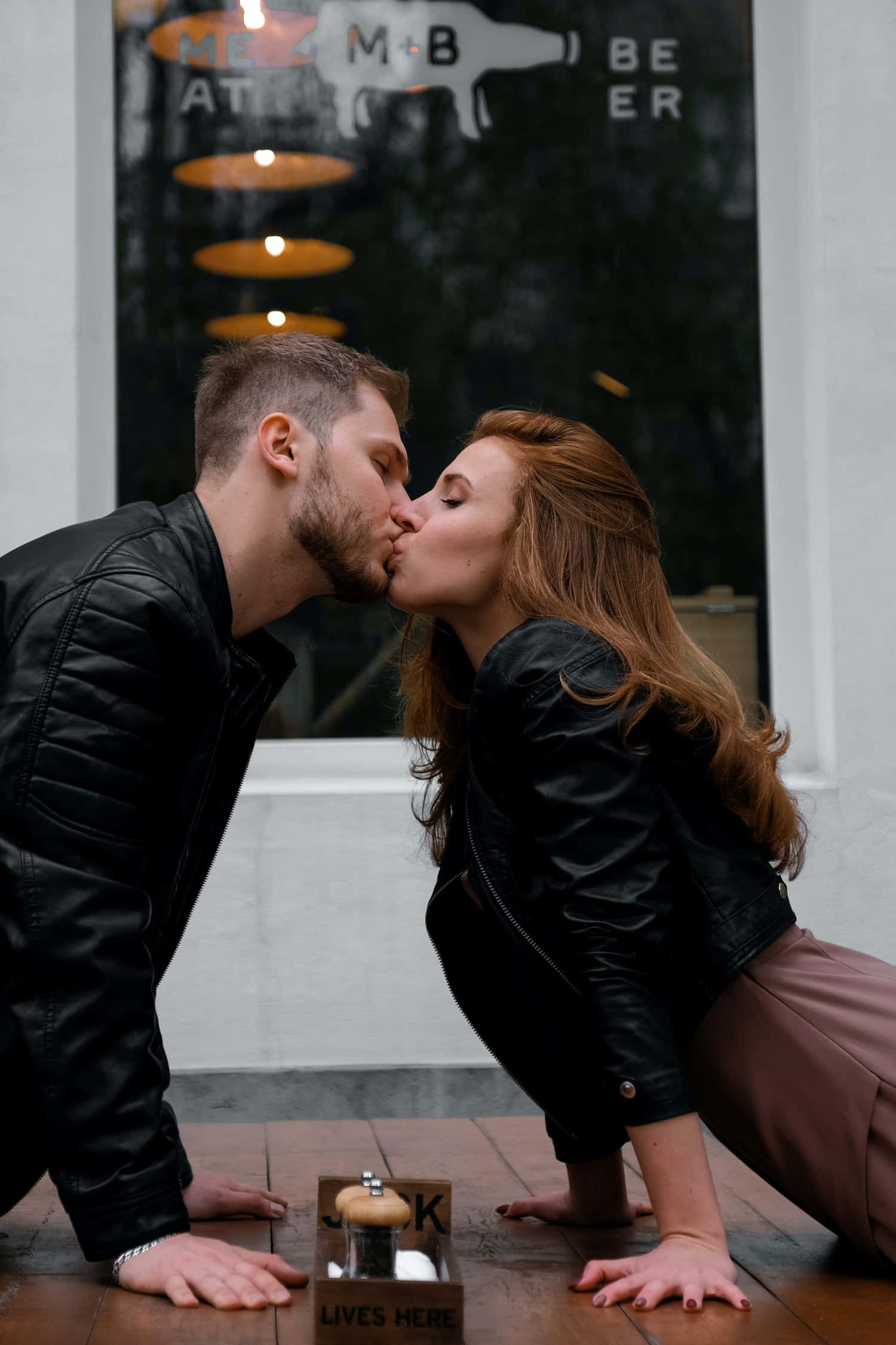 Kissing 桌布 壁紙 — 一對穿著黑色皮夾克的情侶在咖啡館外的木桌上熱情接吻，咖啡館有'MEZ M-B BEER'標誌。