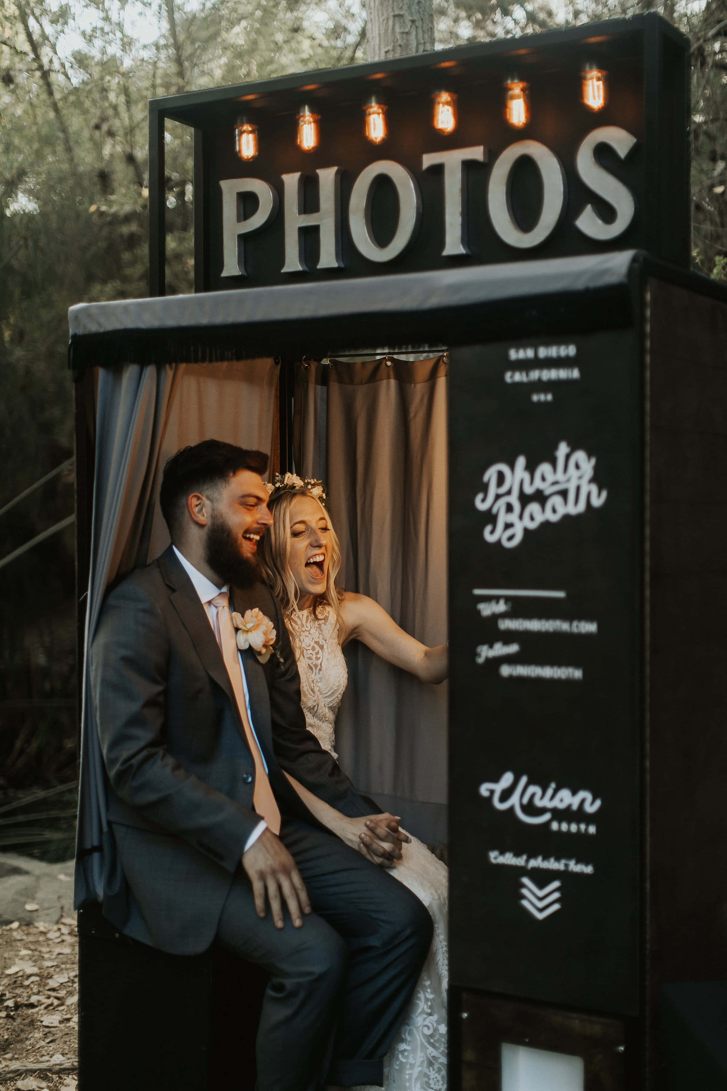 Photo Booth 桌布 壁紙 — 一對笑著的新娘和新郎在樹林地區的溫暖串燈下，於復古風格的拍照亭內開心地擺姿勢。