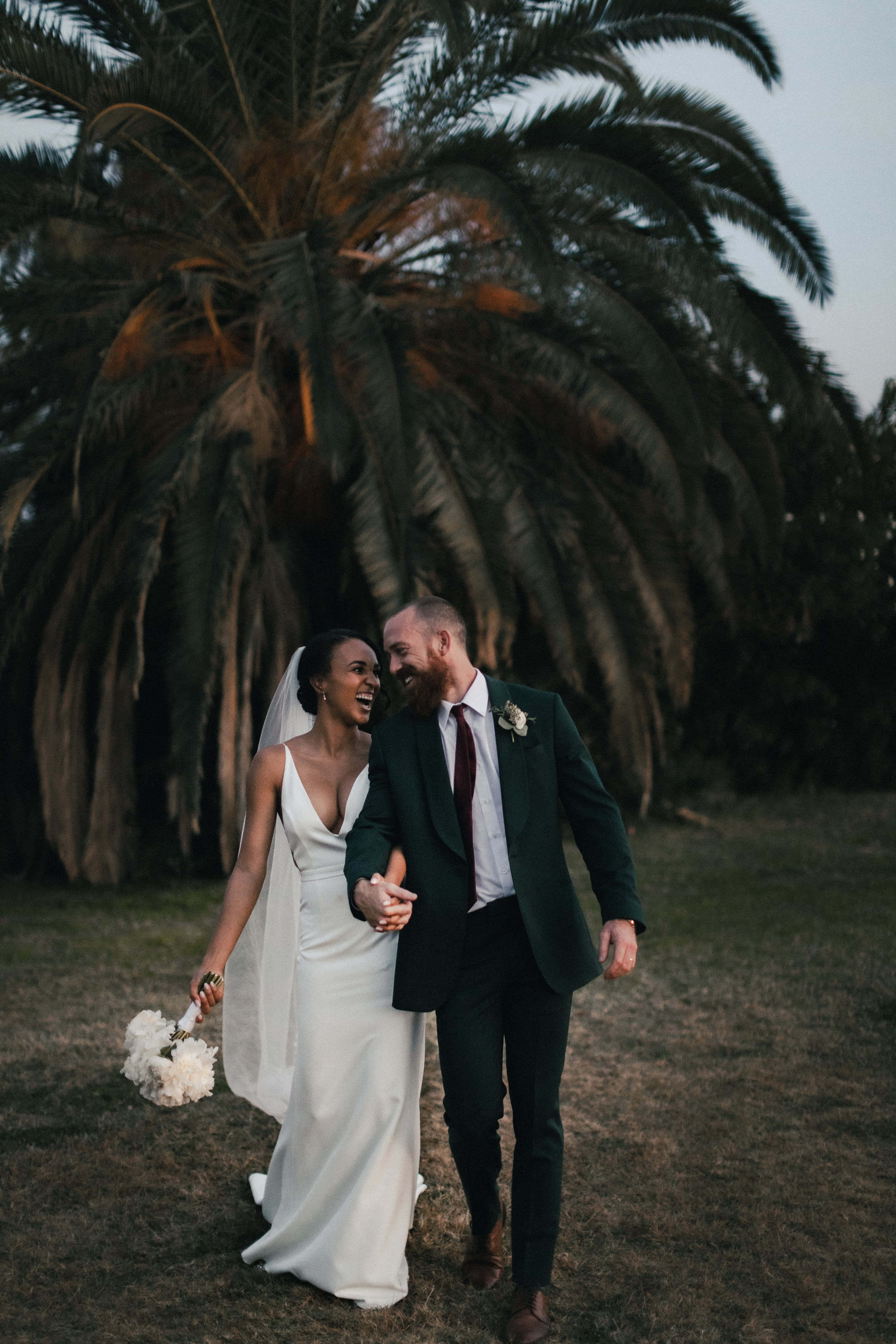 Interracial 桌布 壁紙 — 一對笑著的新娘和新郎手牽手穿過草地，在巨型棕櫚樹下，正值黃金時刻。