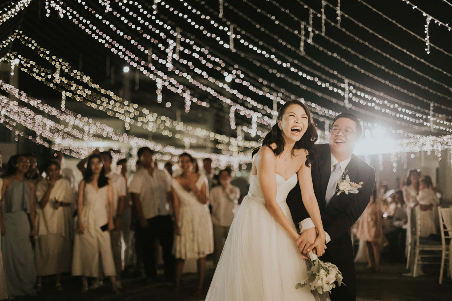 Bride And Groom 桌布 壁紙 — 一對笑著的新郎新娘在閃爍的串燈下於夜間婚禮招待會上分享歡樂時刻。