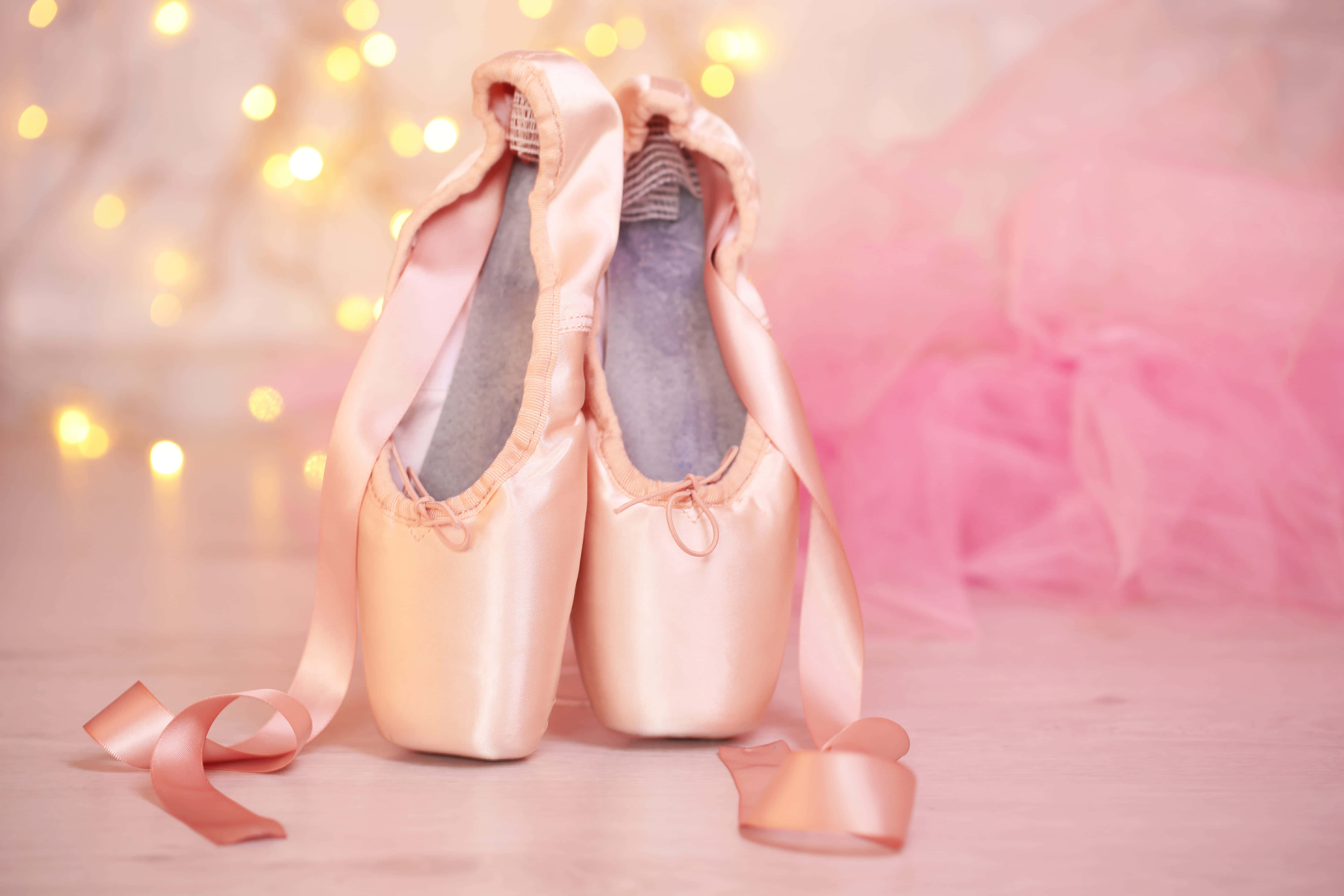 Ballet 桌布 壁紙 — 一對粉紅色緞面芭蕾舞足尖鞋，繫著絲帶，放在柔軟的表面上，旁邊是一件粉紅色芭蕾舞裙，背景有溫暖的散景燈光。