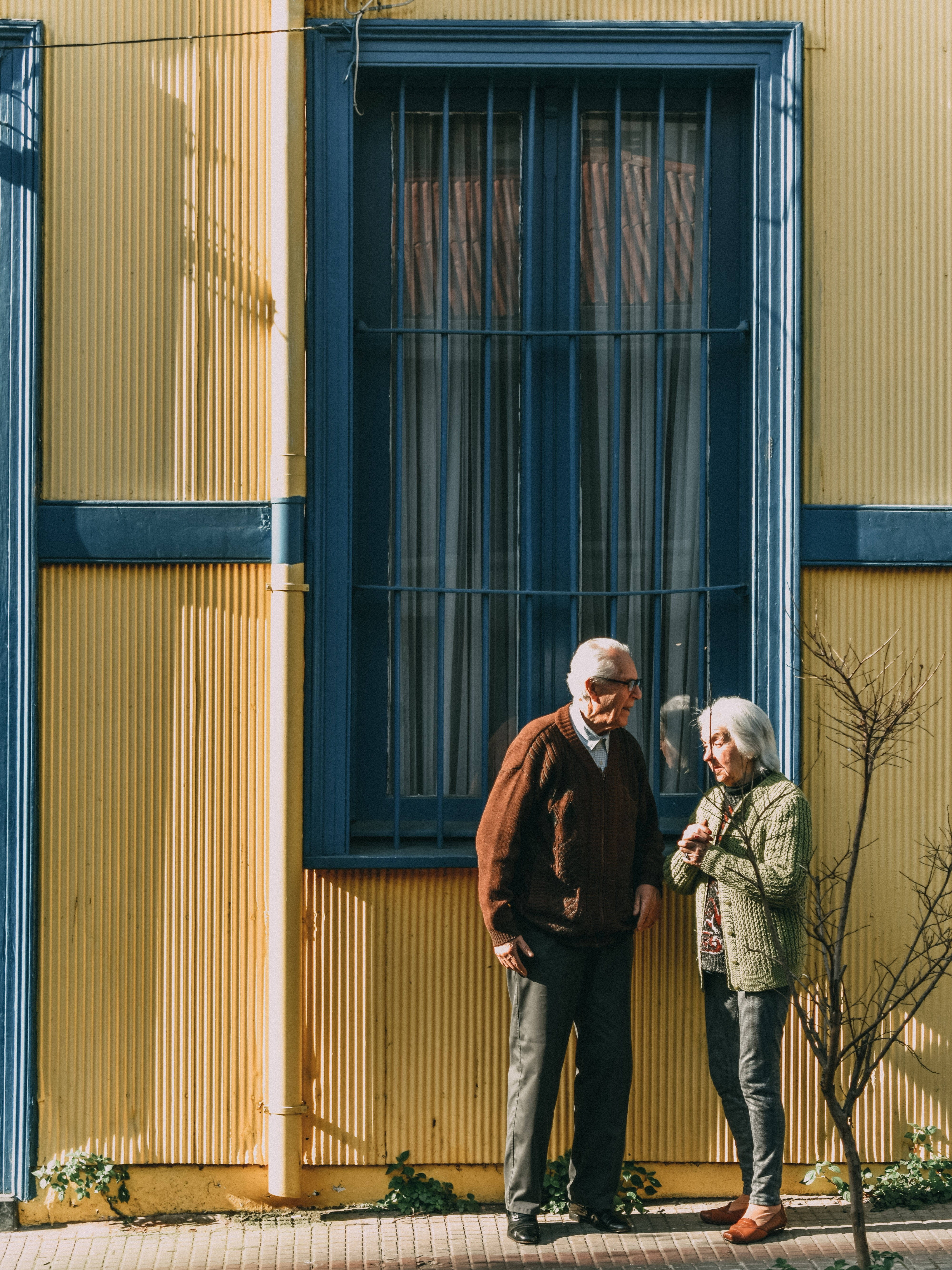 Lovers 桌布 壁紙 — 一對老夫婦站在一棟鮮黃色波紋鐵皮建築旁微笑，建築上有一扇帶欄杆的藍色窗戶。
