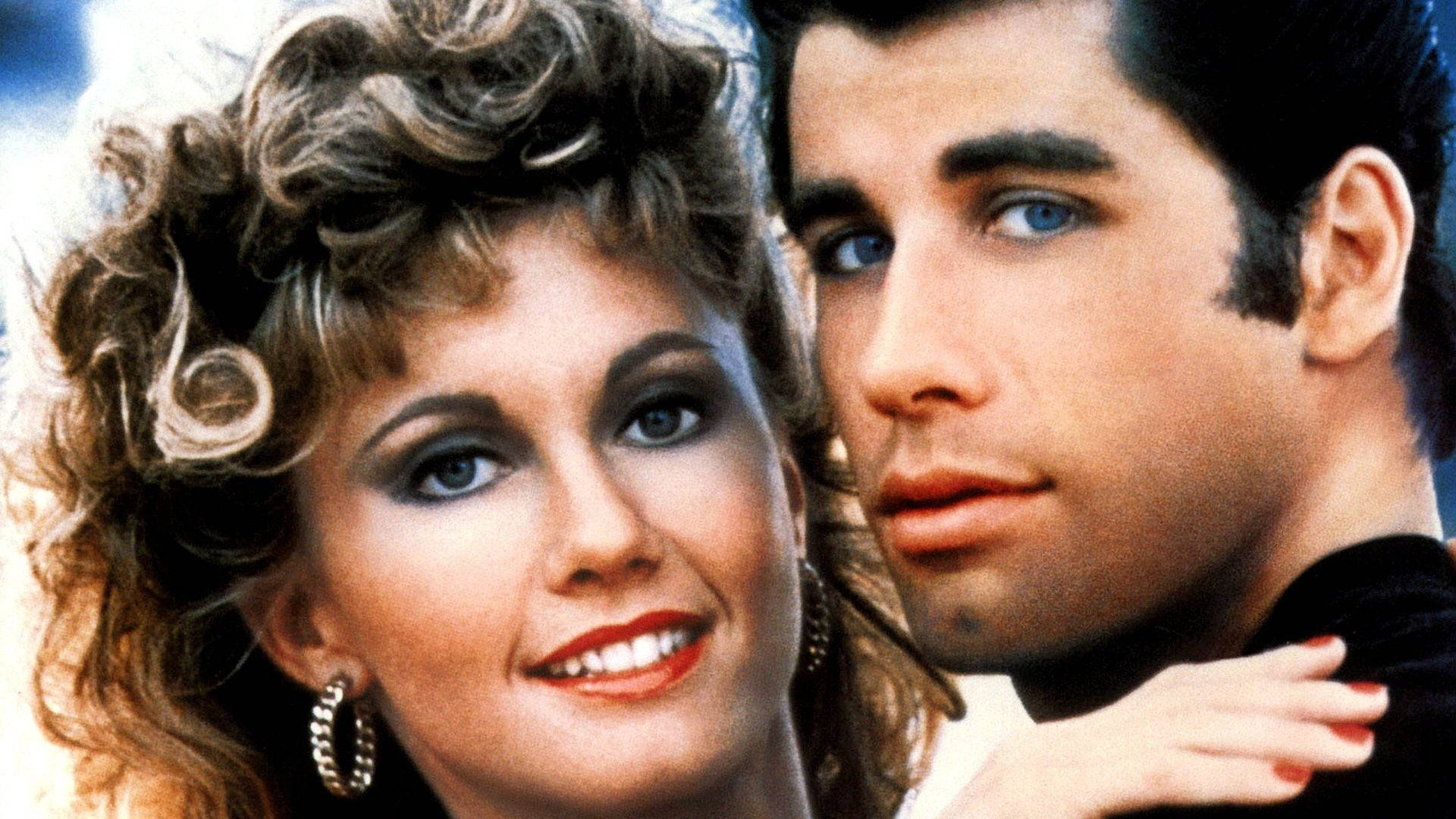 Grease 桌布 壁紙 — 一對華麗的1980年代情侶，擁有豐盈的頭髮和大膽的妝容，在明亮的背景下親密地擺姿勢。