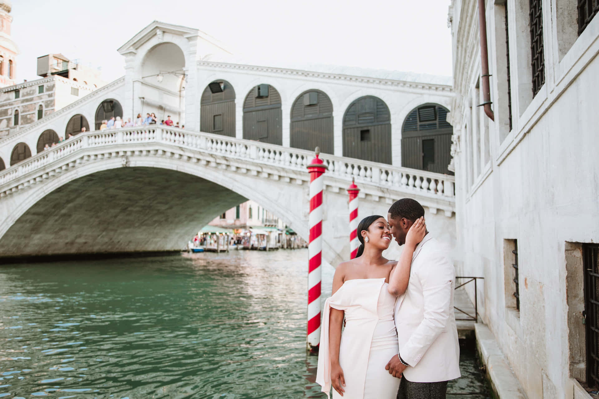 venice 桌布 壁紙 — 一對身穿白色婚紗的情侶在威尼斯標誌性的里亞爾托橋下的運河旁分享親密時刻。