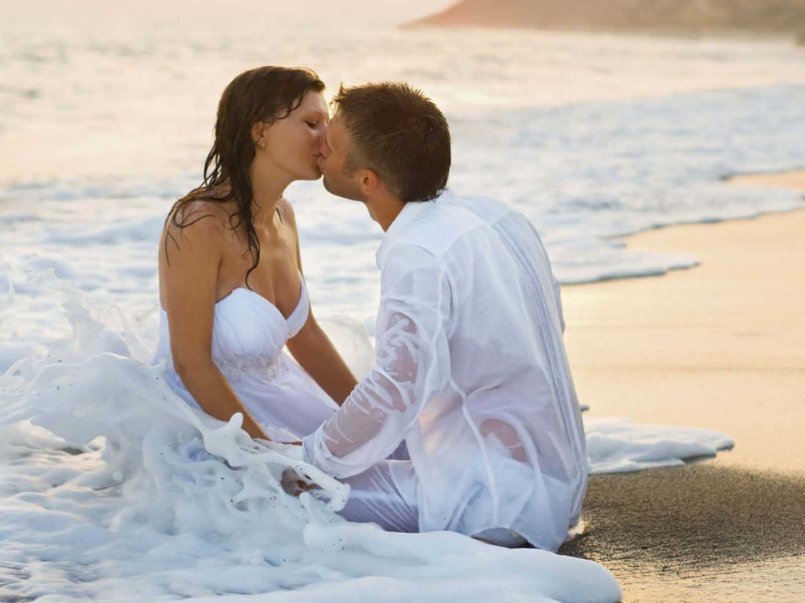 Beach Wedding 桌布 壁紙 — 一對身穿白色婚紗的情侶在陽光明媚的海灘上熱情接吻，海浪在他們周圍拍打。