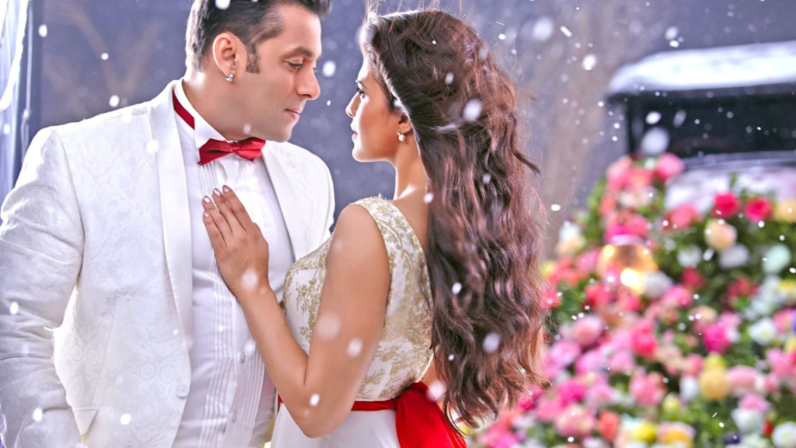 Salman Khan 桌布 壁紙 — 一對身穿白色正裝的情侶在飄雪下分享親密時刻，附近有色彩繽紛的花藝佈置。