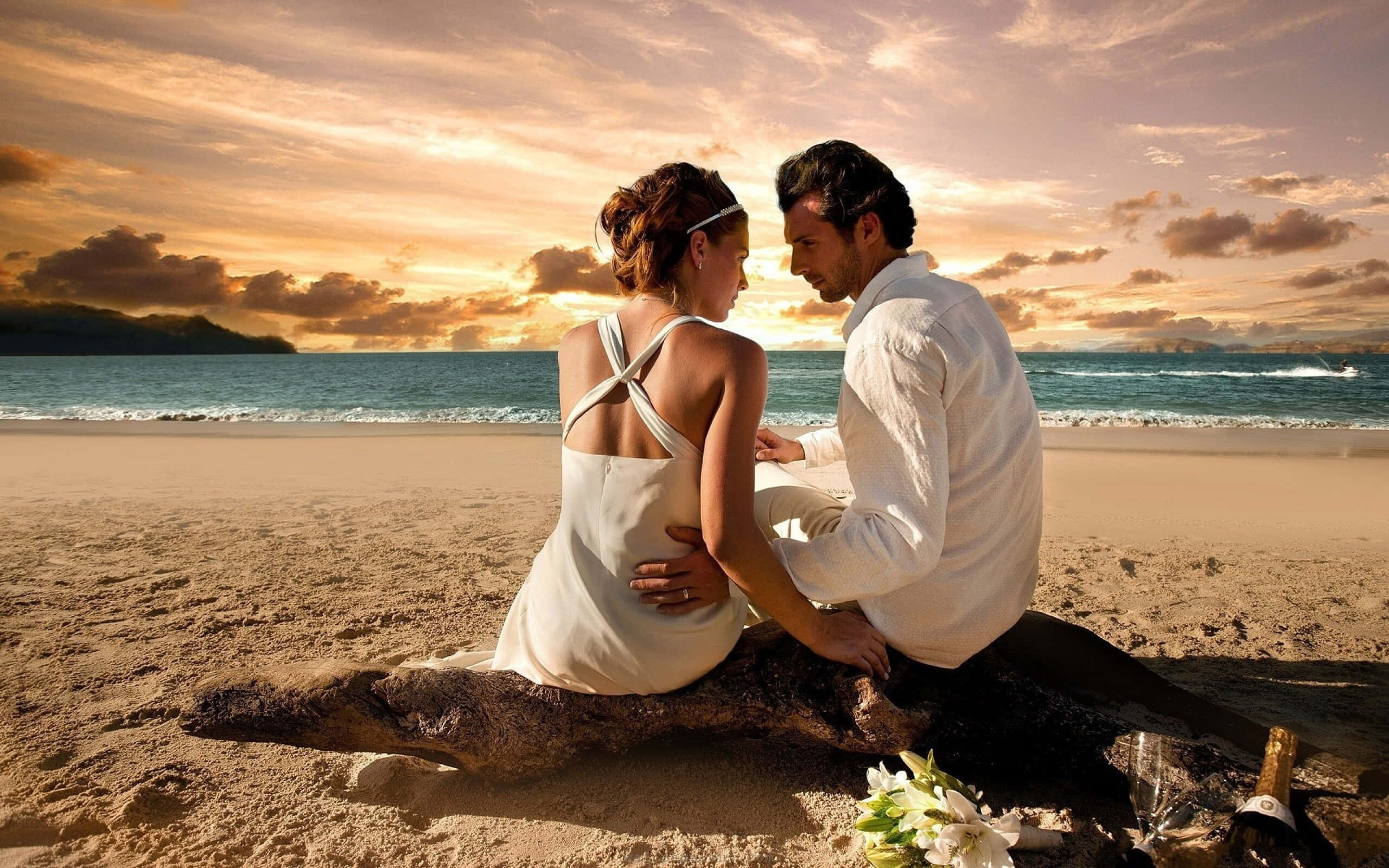 Beach Lovers 桌布 壁紙 — 一對身穿白衣的情侶在日落時分親密地坐在沙灘的浮木上，海面波濤起伏，天空金黃燦爛。