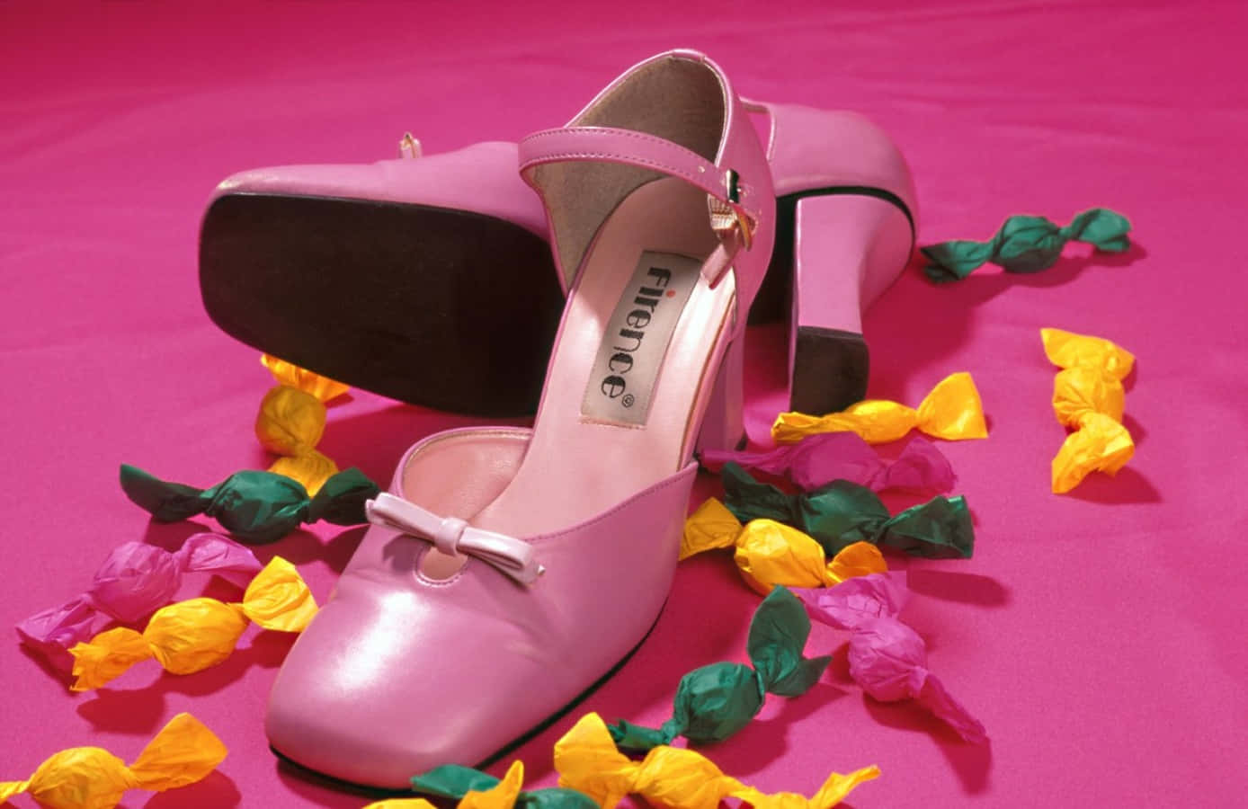 Pink Shoes 桌布 壁紙 — 一對閃亮的粉紅色高跟鞋，帶有蝴蝶結，放在充滿活力的粉紅色表面上，周圍環繞著色彩繽紛的包裝糖果。