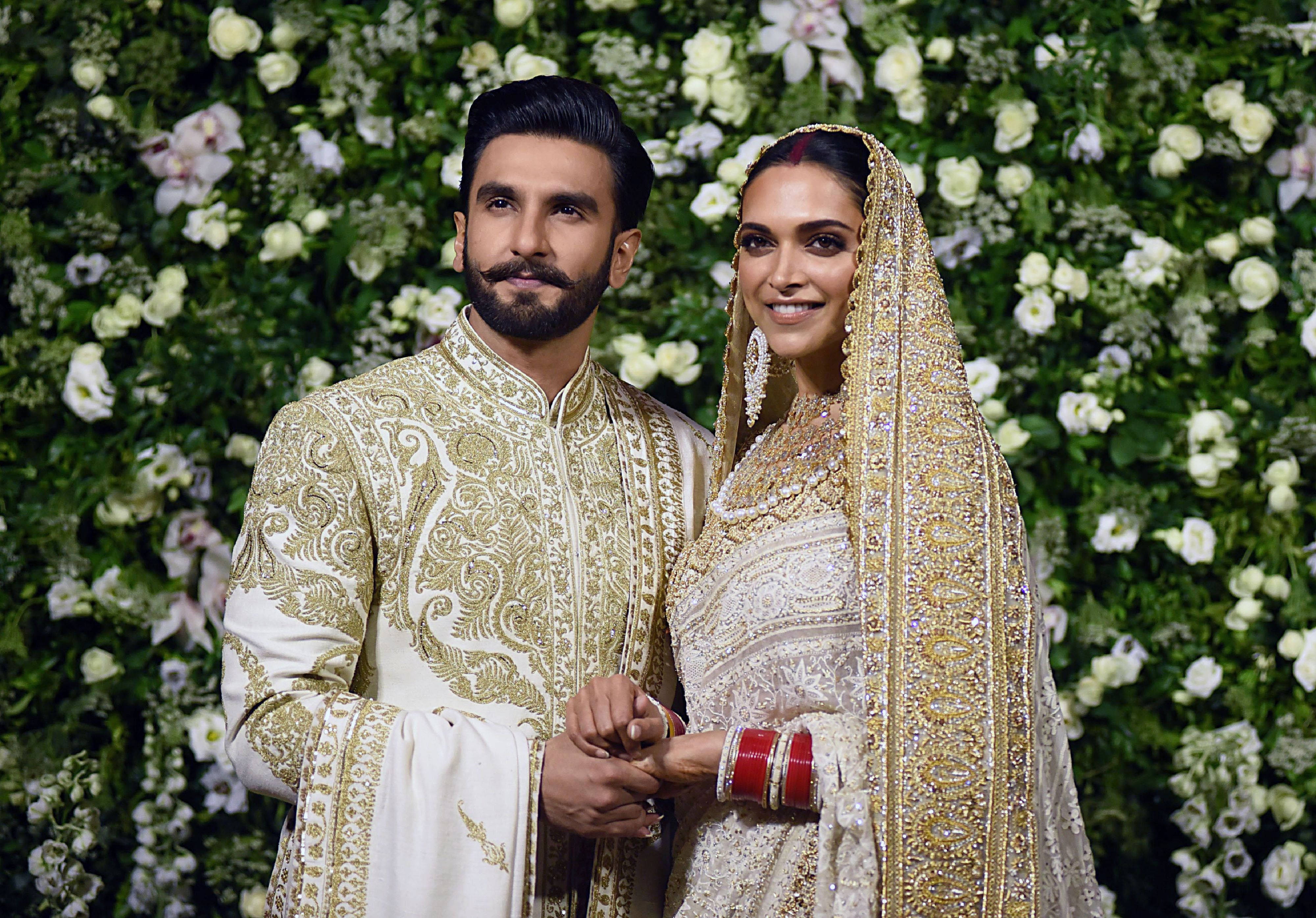 Ranveer Singh 桌布 壁紙 — 一對閃耀的新人穿著華麗的白色和金色婚禮服飾，在茂盛的花卉背景前牽手。