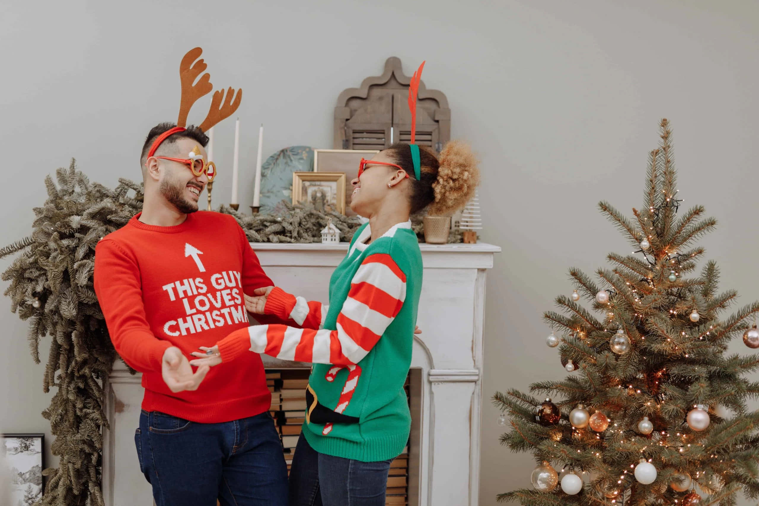 Couples Christmas 桌布 壁紙 — 一對開心的情侶戴著節日馴鹿角及聖誕毛衣，在裝飾好的聖誕樹旁一起大笑。