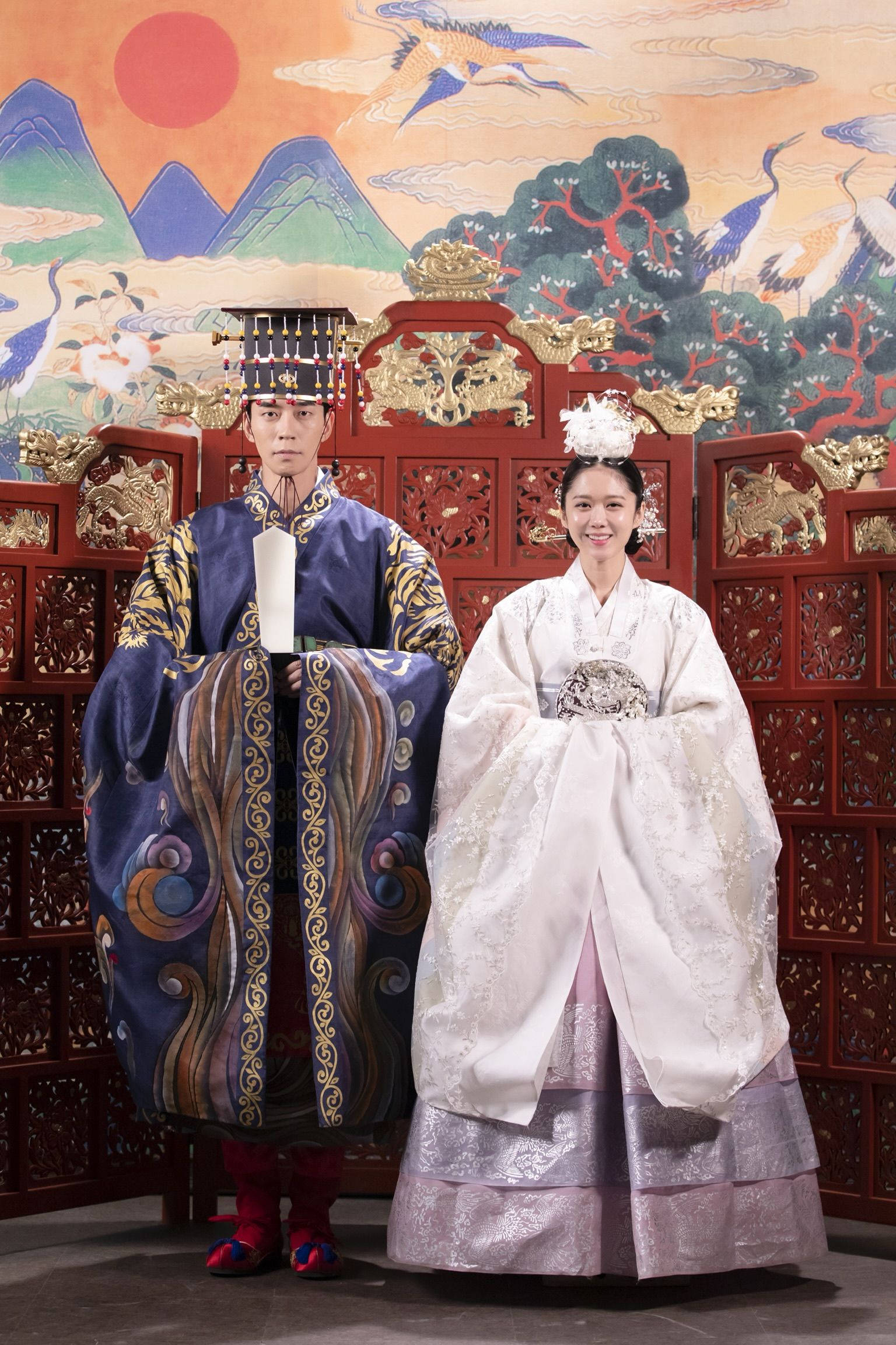 kdrama 桌布 壁紙 — 一對韓國新人穿著傳統婚禮韓服，站在華麗的紅色屏風前，屏風上繪有山脈和鶴，背景是紅色的太陽。