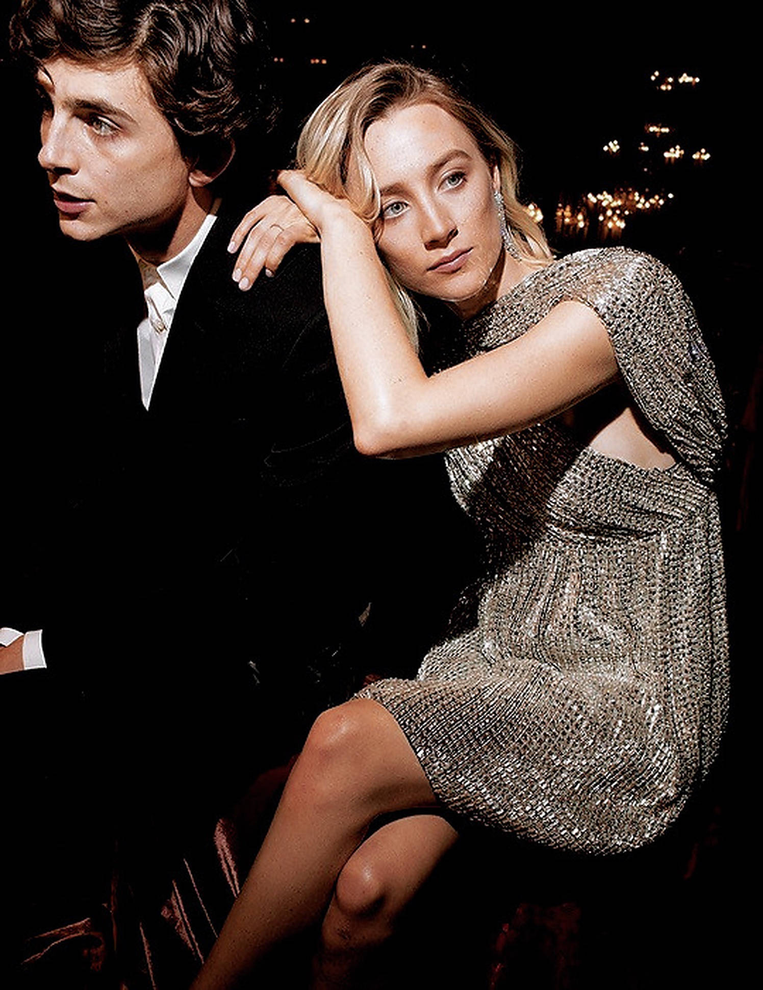 Saoirse Ronan 桌布 壁紙 — 一對魅力情侶在夜晚擺姿勢，背後城市燈光閃爍，身穿閃亮銀色連衣裙的女子直視鏡頭。